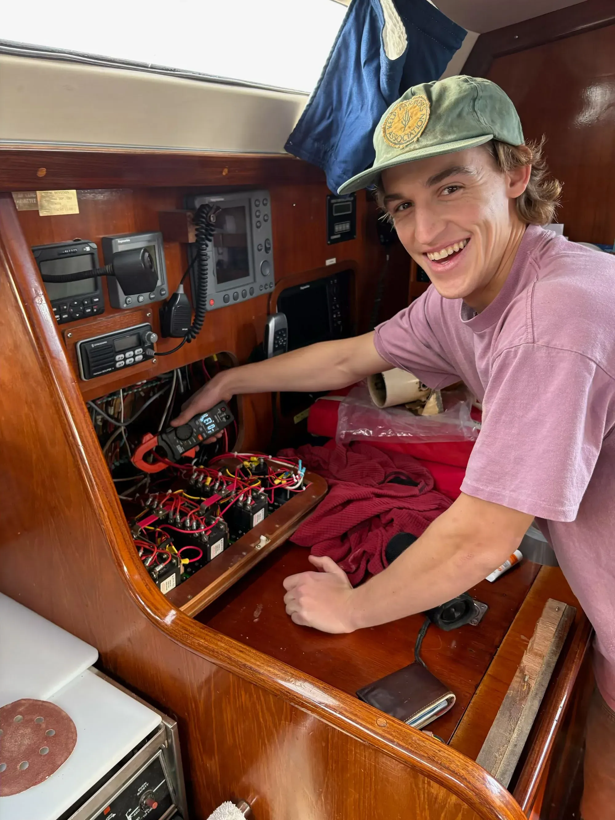 شاب مبتسم يقوم بفحص الأسلاك الكهربائية داخل قارب شراعي، مع وجود معدات بحرية وأدوات في الخلفية، مما يعكس روح المغامرة والإبحار.