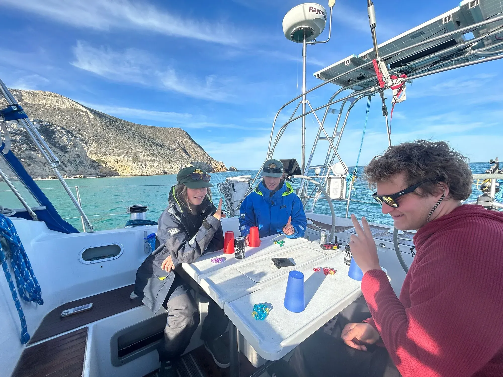 مجموعة من الأصدقاء يلعبون ألعاب الطاولة على متن قارب شراعي، مع منظر لجبل خلفهم ومياه المحيط الهادئ.