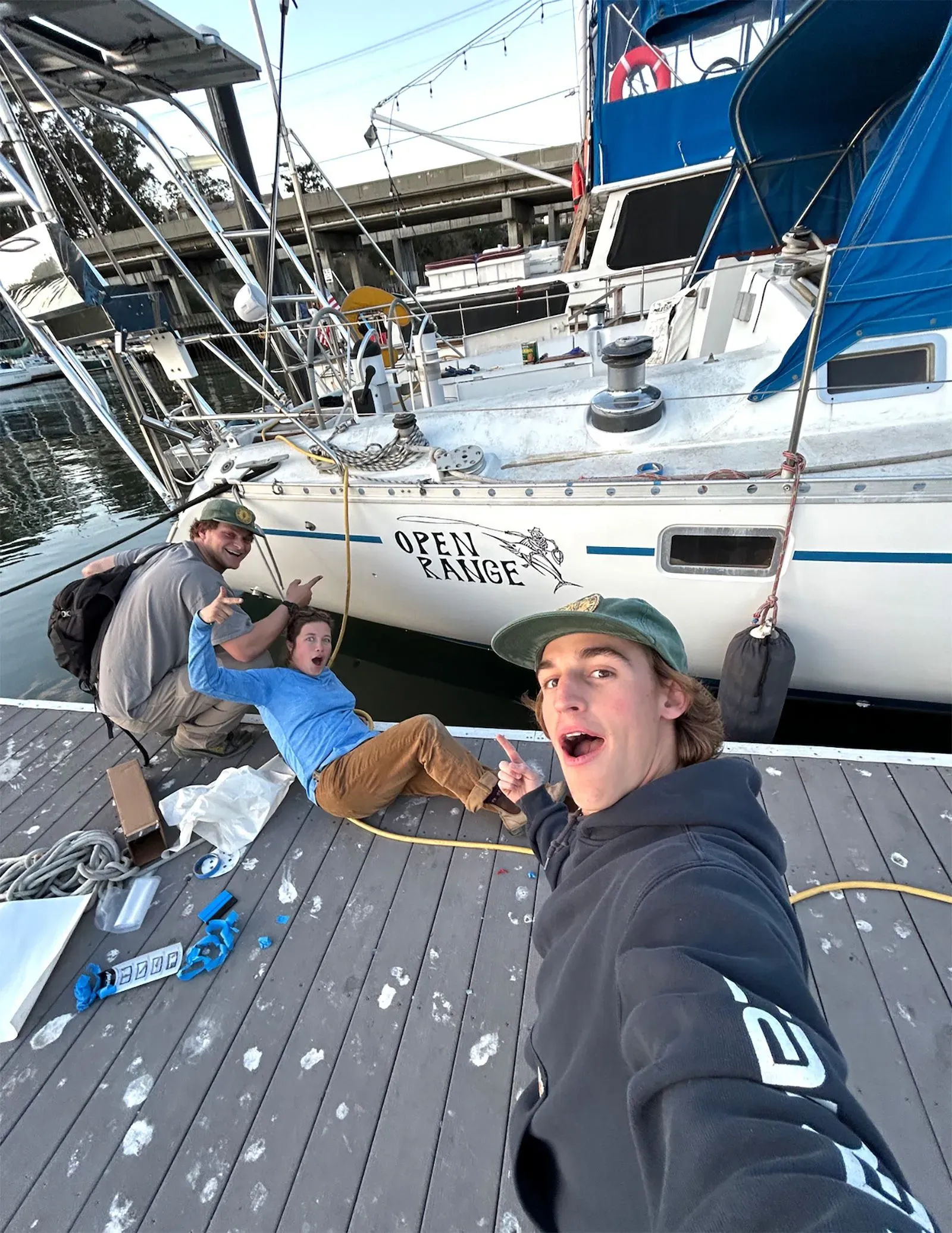مجموعة من الأصدقاء يقفون بجوار قارب شراعي يحمل اسم "OPEN RANGE" في المرسى، مع تعبيرات حماسية استعدادًا لمغامرتهم في المحيط.