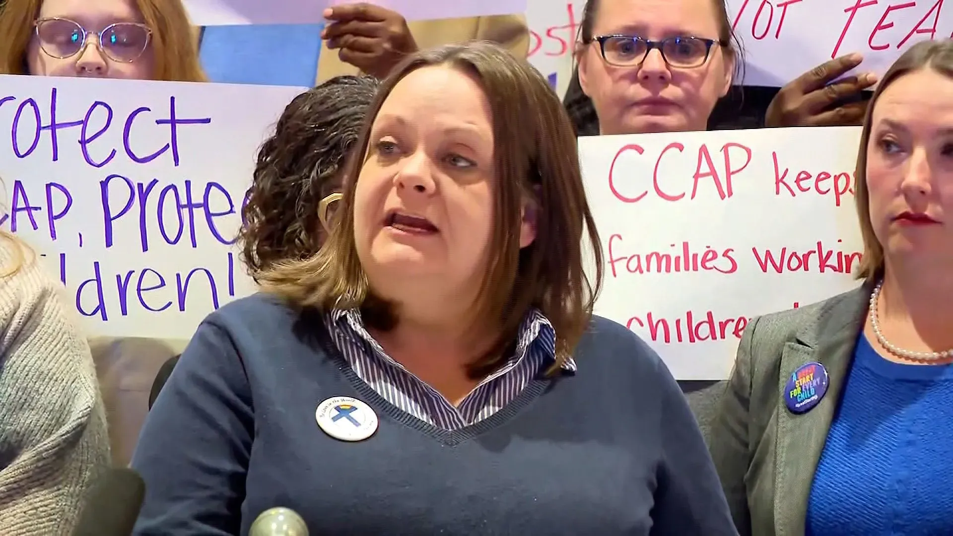 امرأة تتحدث في مؤتمر صحفي حول تأثير تجميد تمويل رعاية الأطفال في مينيسوتا، بينما يحمل الحضور لافتات تدعم الأسر.