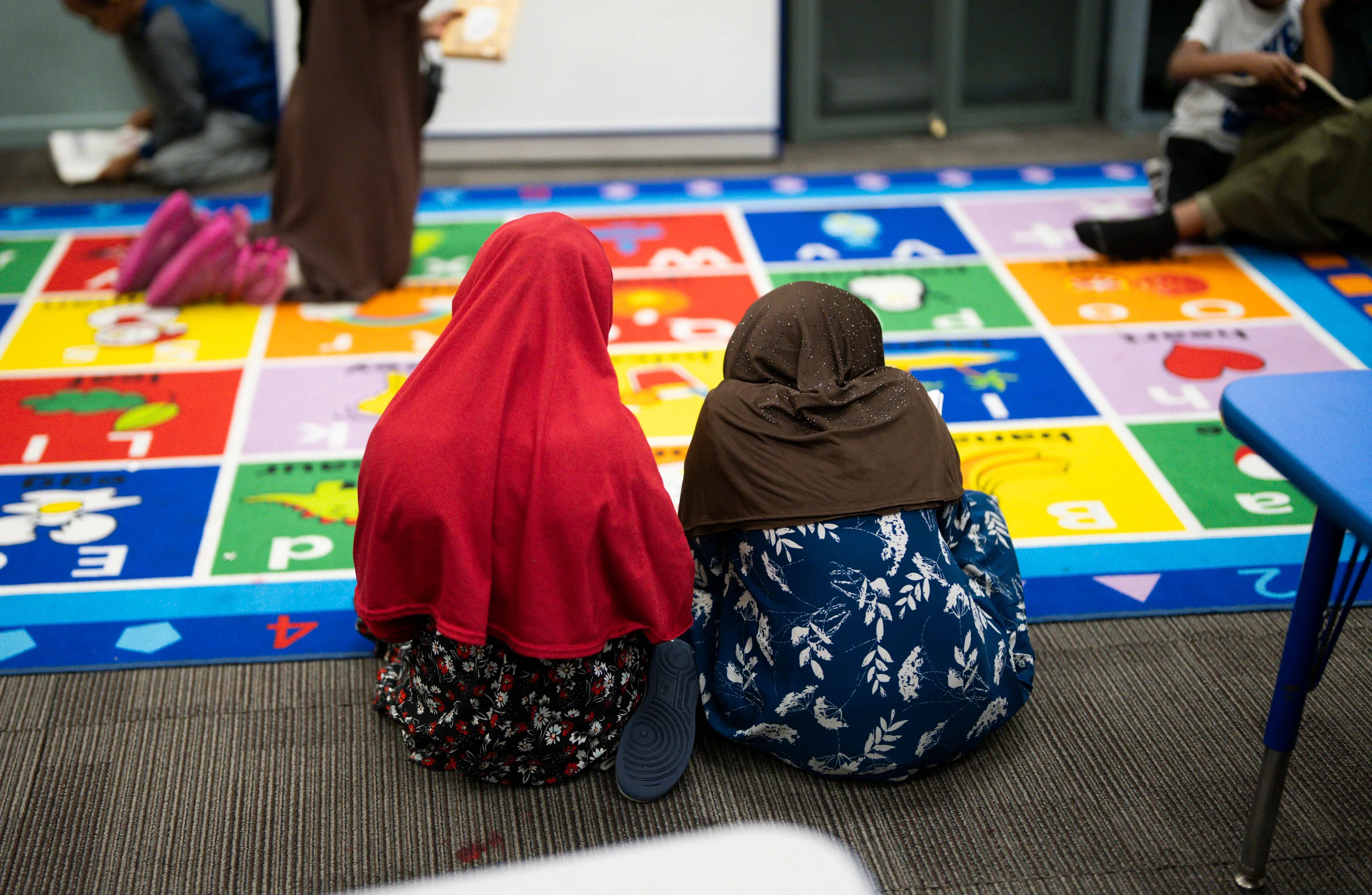 طفلتان ترتديان الحجاب تجلسان على سجادة ملونة في مركز رعاية الأطفال، مع أطفال آخرين مشغولين بالأنشطة.