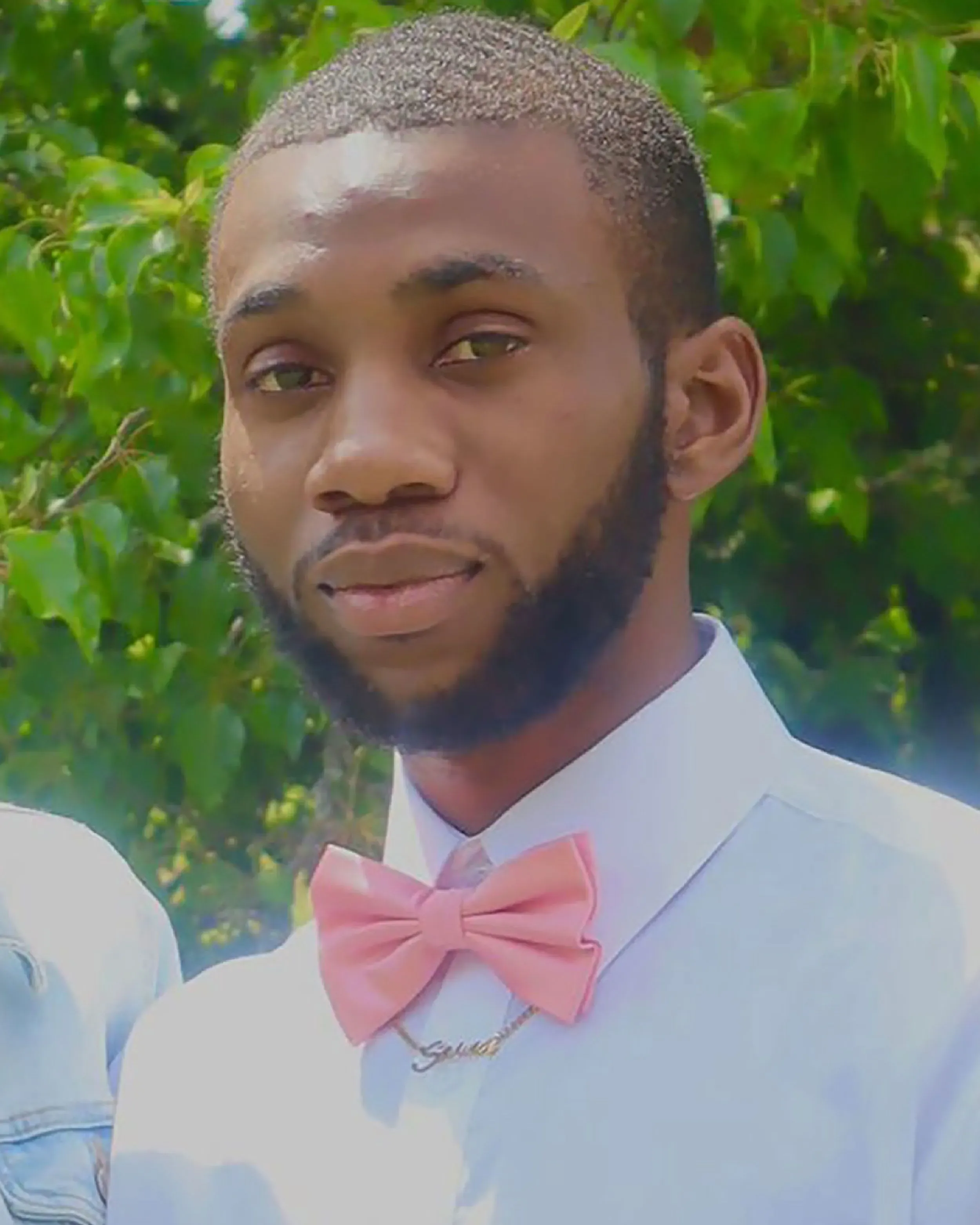 صورة شاب يرتدي قميصًا أبيض وربطة عنق وردية، يظهر بابتسامة خفيفة، مع خلفية من الأشجار الخضراء، تعبيرًا عن الحياة والأمل.