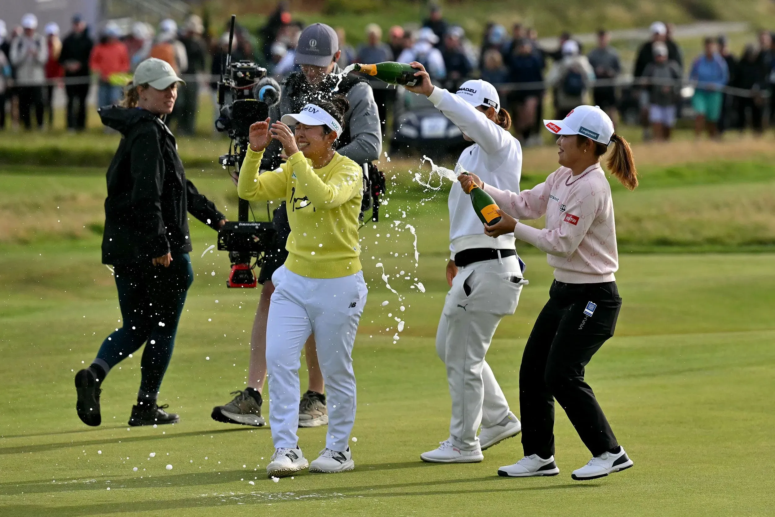 ميو ياماشيتا تحتفل بفوزها في بطولة AIG المفتوحة للسيدات، محاطة بزميلاتها وهن يسكبن الشمبانيا، مع جمهور كبير في الخلفية.