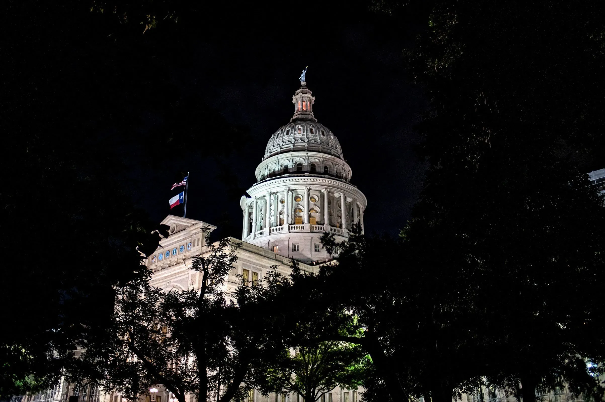 مبنى الكابيتول في تكساس مضاء ليلاً، مع العلم الأمريكي يرفرف فوق القبة، محاط بأشجار خضراء، مما يعكس أهمية السياسة المحلية.