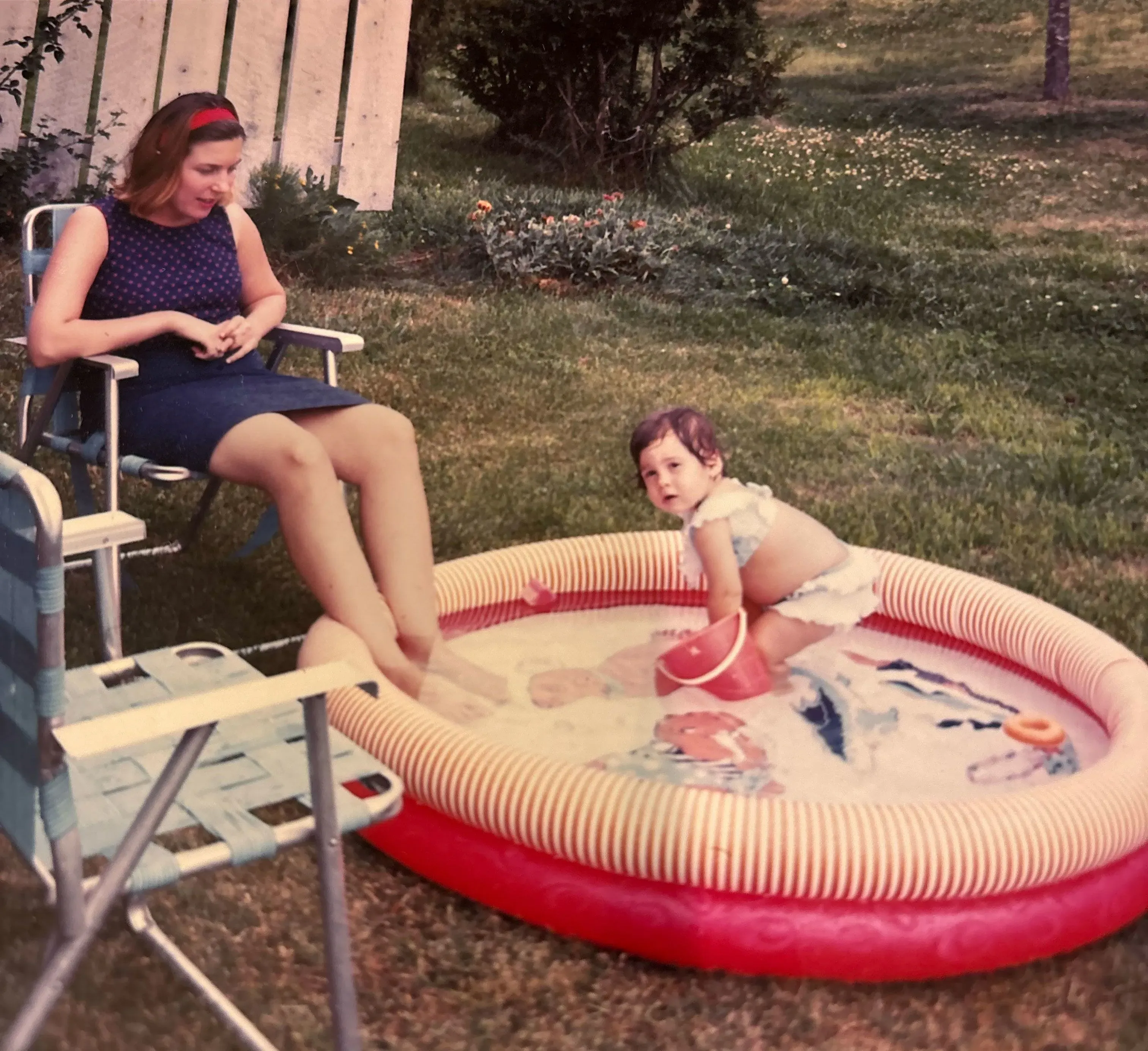 امرأة تجلس على كرسي بجانب بركة سباحة صغيرة، بينما طفل يلعب داخل البركة. تعكس الصورة لحظات عائلية دافئة في الطبيعة.