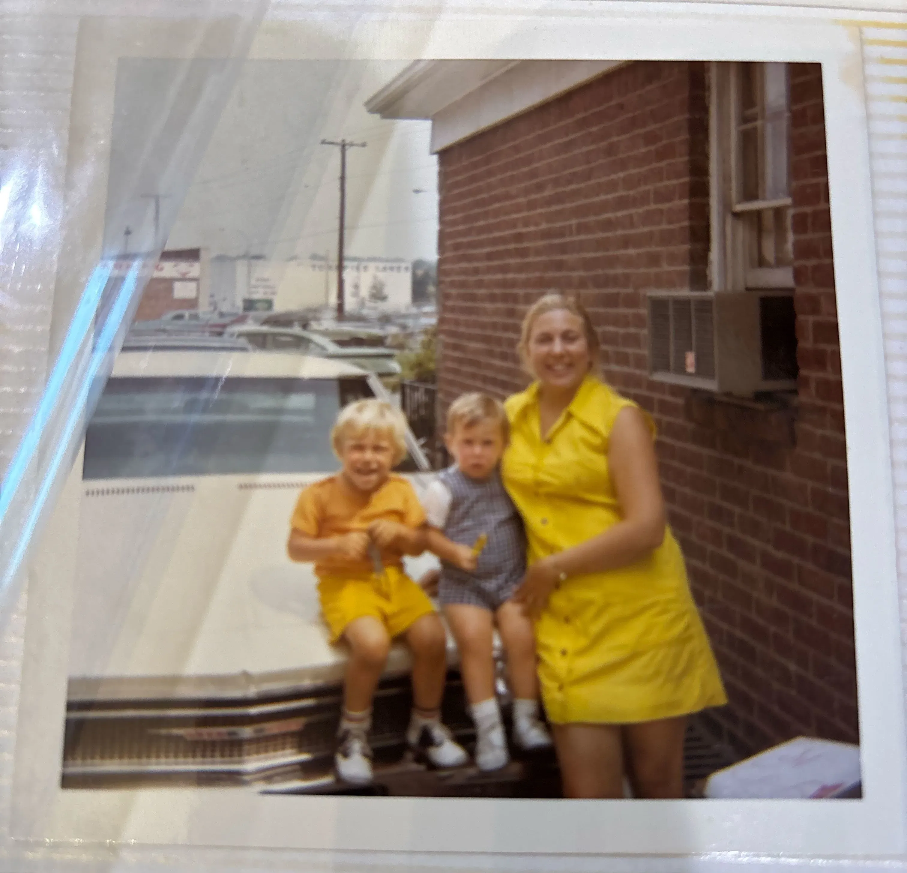 صورة عائلية تظهر امرأة ترتدي فستانًا أصفر مع طفلين يجلسان على مقدمة سيارة، تعكس لحظة من السعادة في حياة مارلا والدمان قبل مأساة فقدان والدتها.