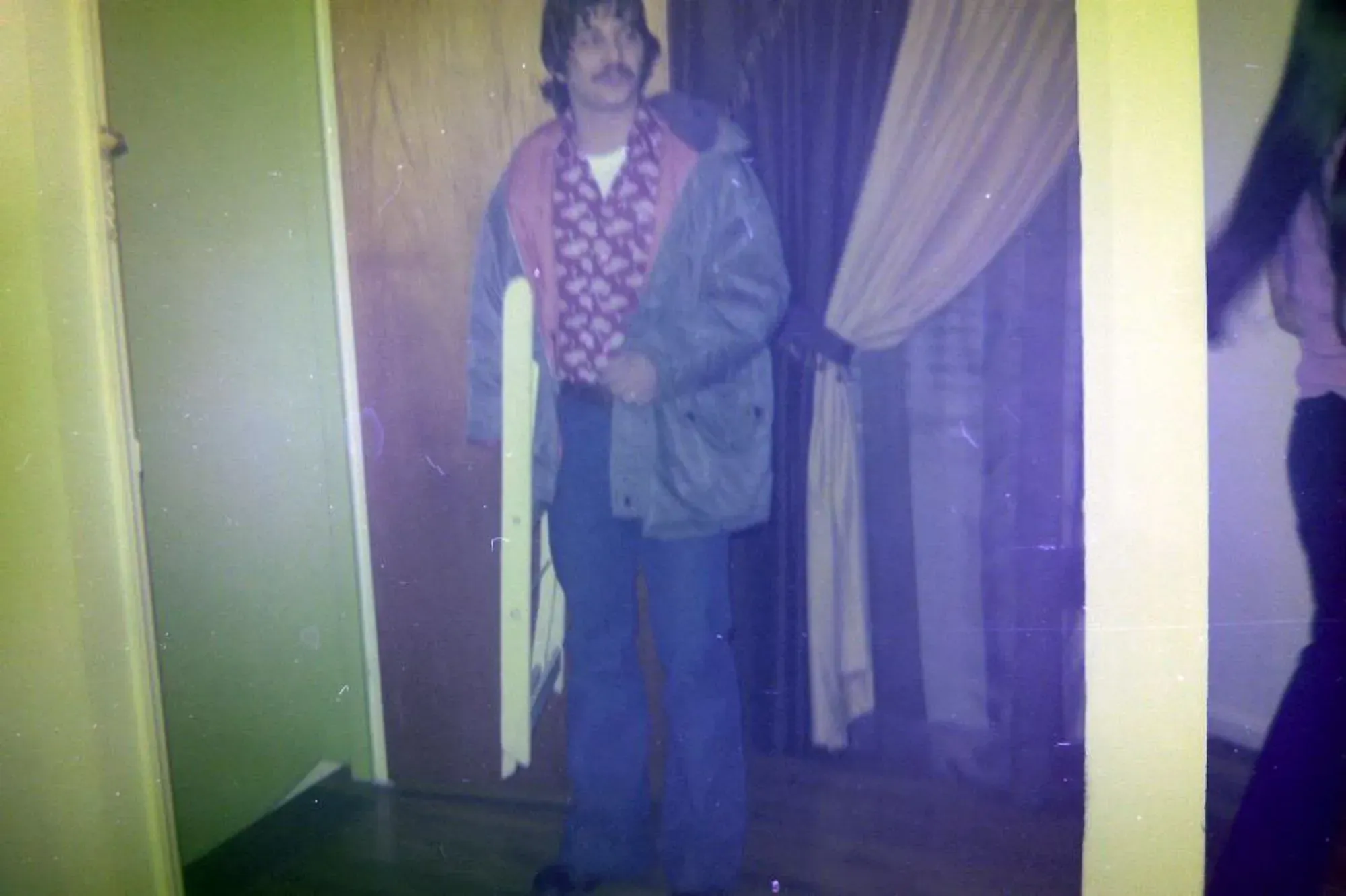 صورة لرجل يرتدي معطفًا ثقيلًا وقميصًا مزخرفًا، يقف في مدخل غرفة ذات جدران صفراء، مما يثير التساؤلات حول دوره في قضية قتل قديمة.