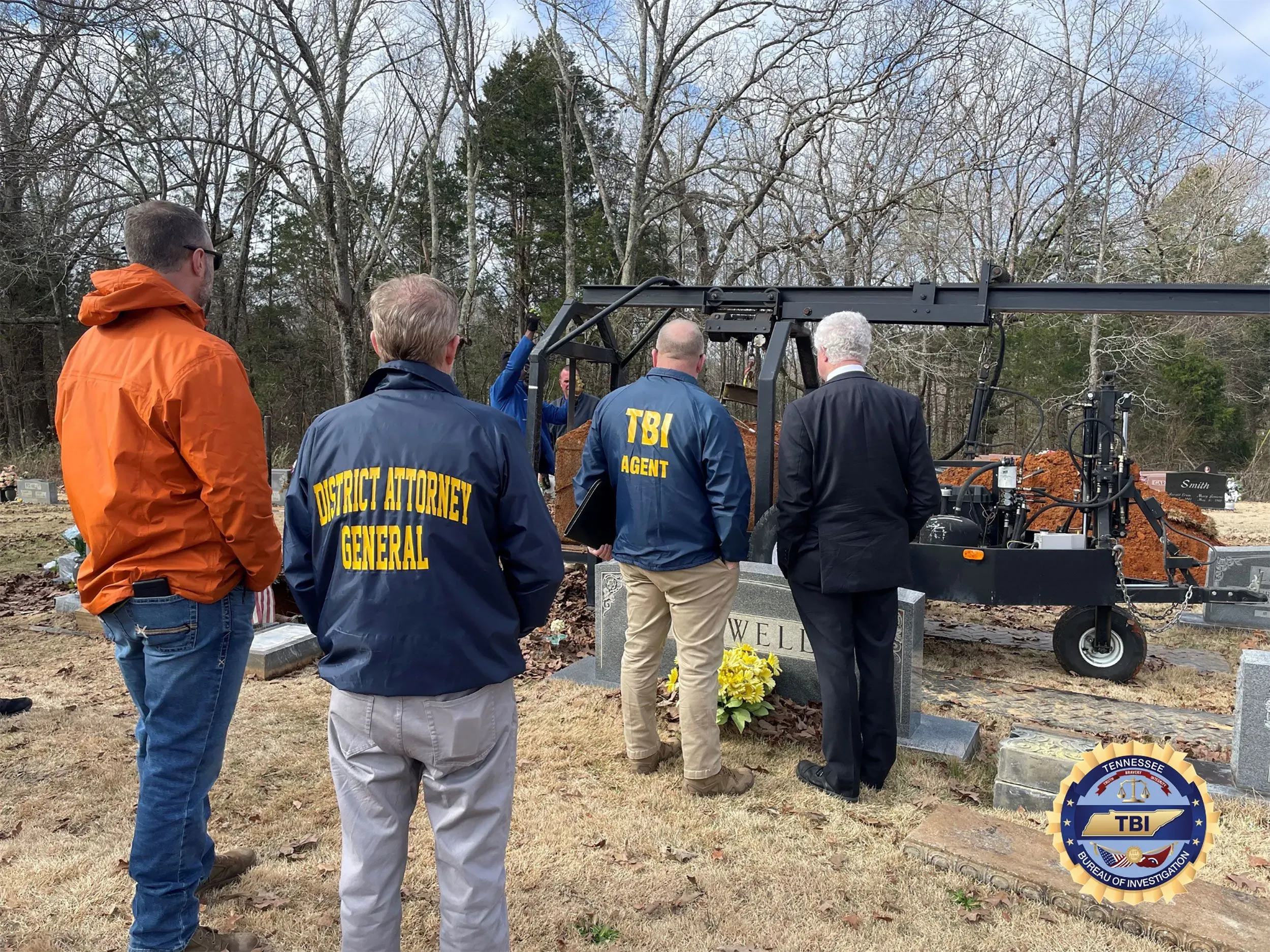 مجموعة من المحققين والمسؤولين يقفون أمام قبر في مقبرة، حيث يتم استخدام معدات لرفع التربة، مع وجود زهور بجانب القبر.