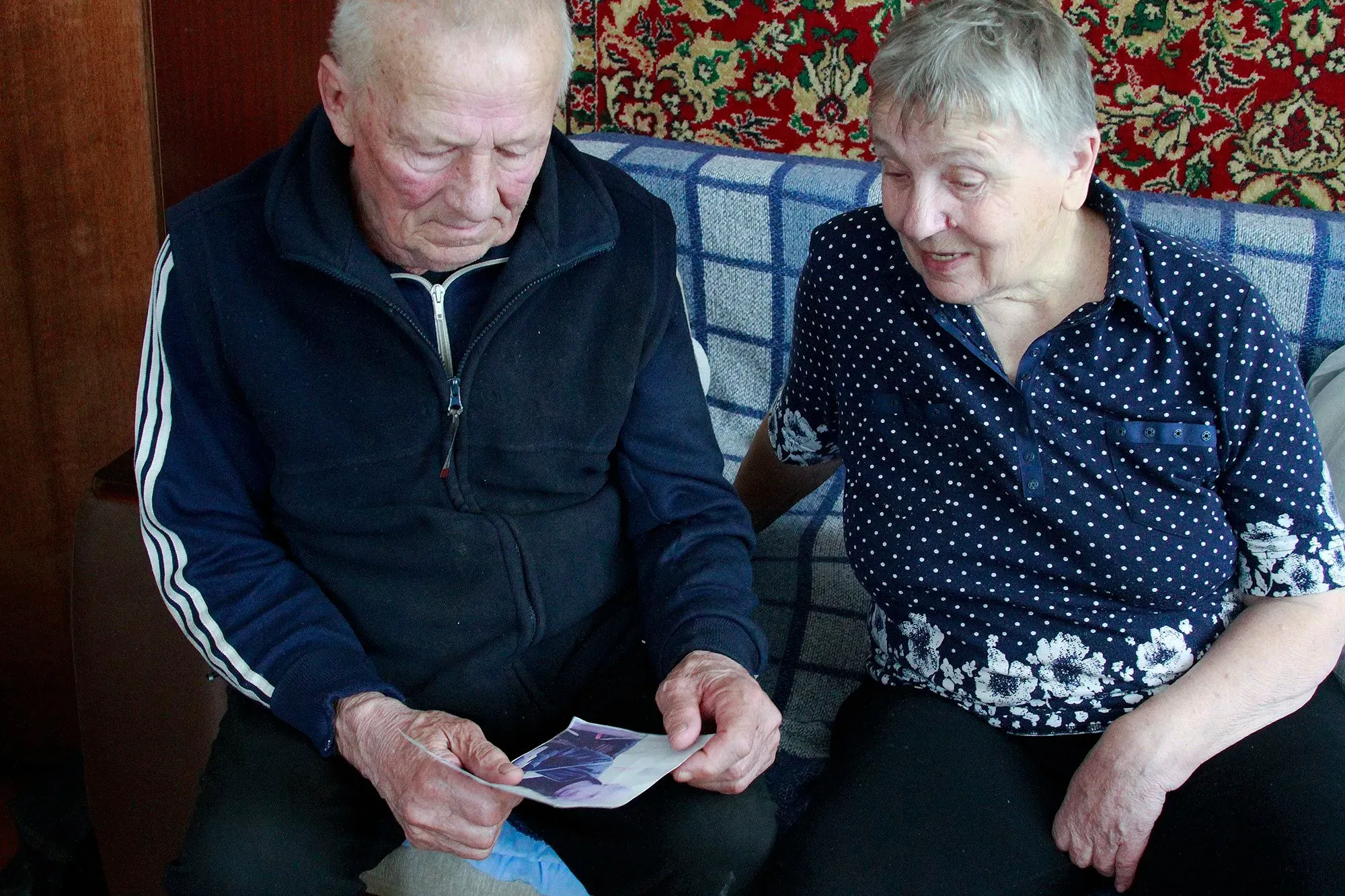 زوجان مسنّان يجلسان معًا في غرفة معيشتهما، يتبادلان نظرات الأمل وهما ينظران إلى صورة، تعبيراتهما تعكس مشاعر الفرح والحنين.