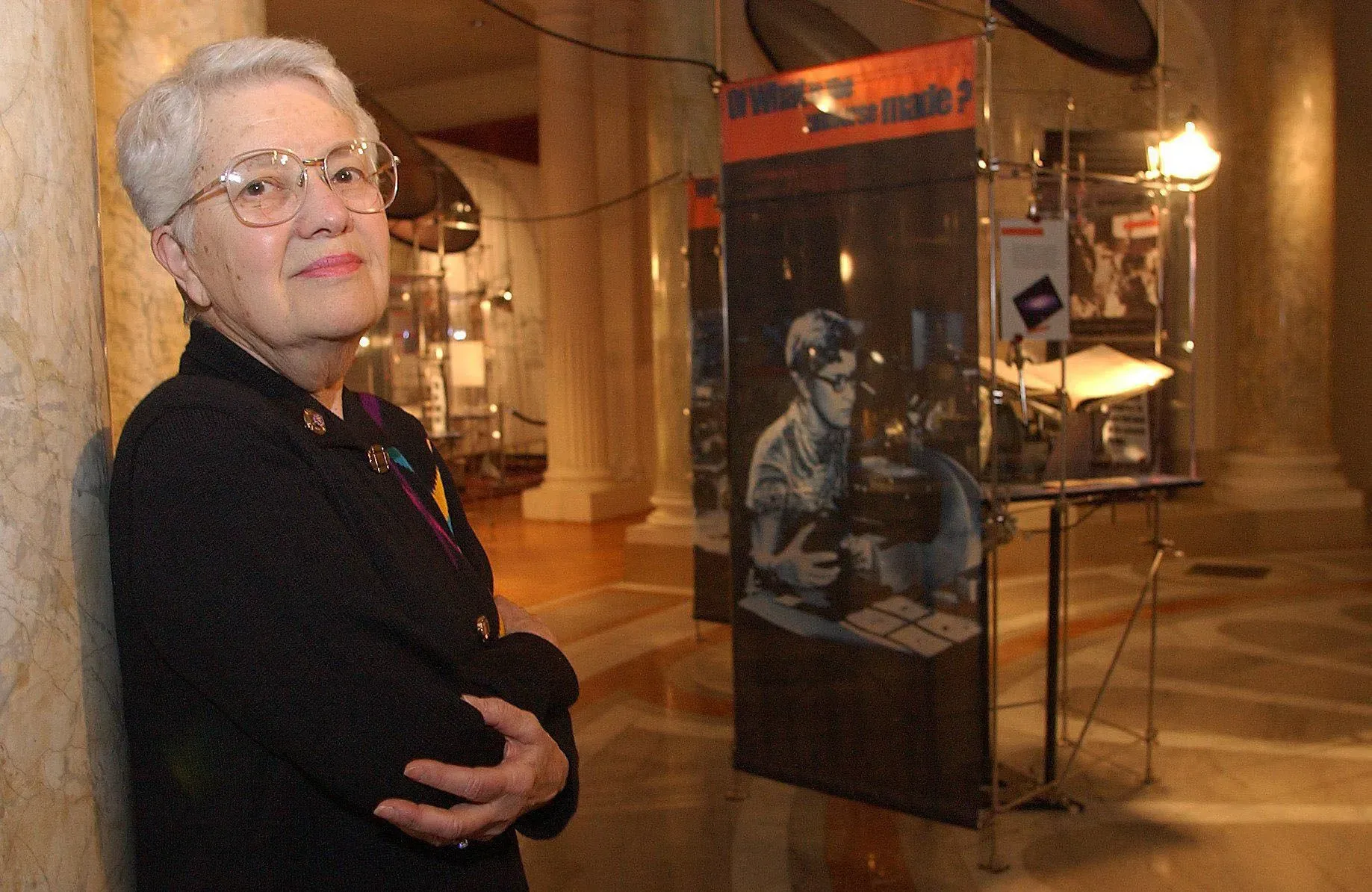 امرأة مسنّة ترتدي نظارات وتبتسم، تقف بجوار معرض علمي في متحف، مع لافتات توضح تجارب علمية وأبحاث.