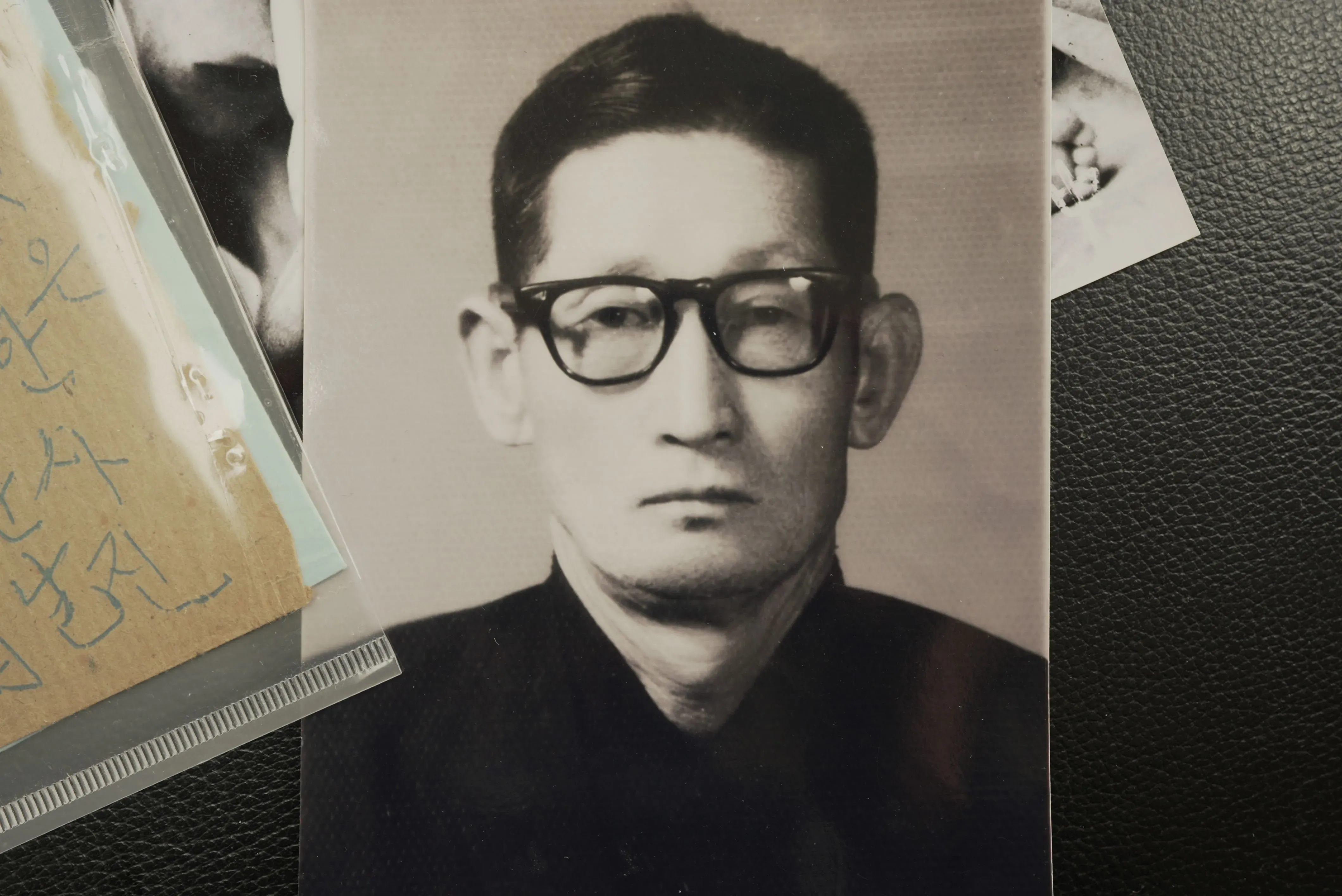 صورة بالأبيض والأسود لرجل يرتدي نظارات، يظهر بملامح جدية، محاطًا بمستندات وصور أخرى، تعكس تاريخ اللاجئين الكوريين.
