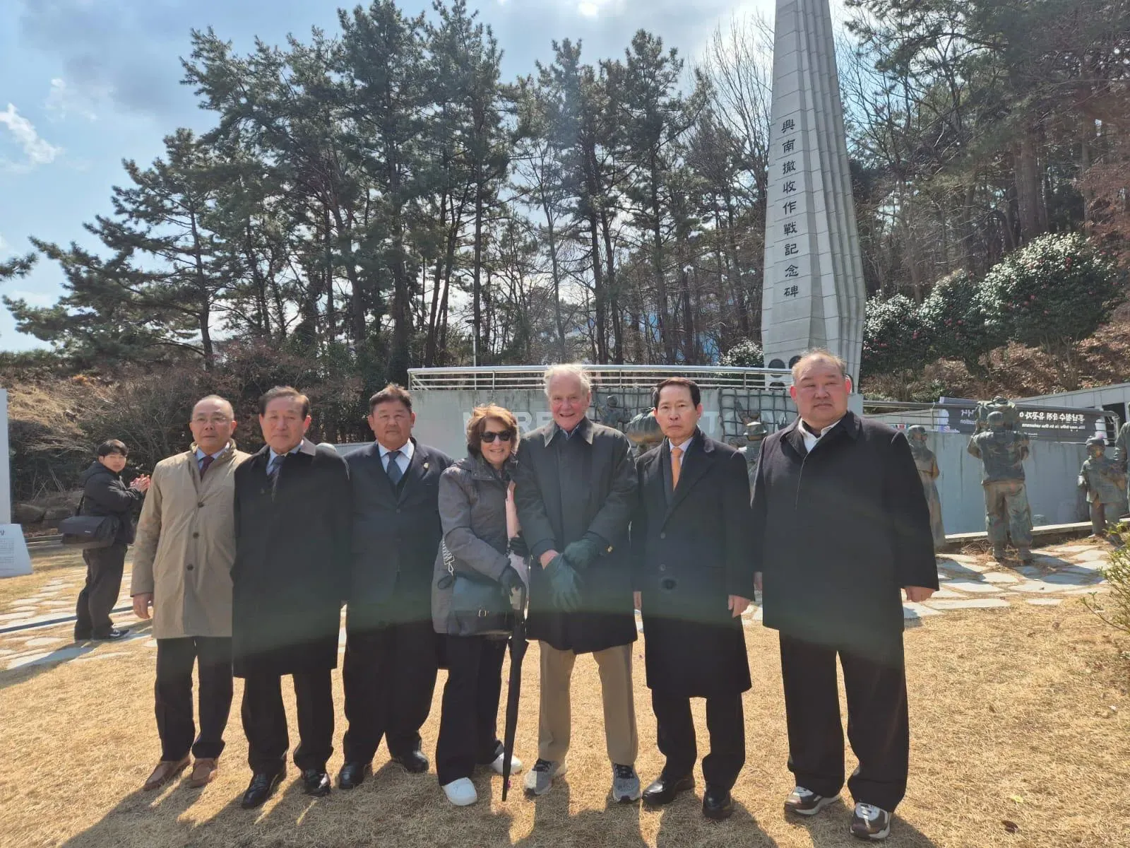 تجمع مجموعة من الأشخاص أمام نصب تذكاري، مع أشجار خلفية، في ذكرى أحداث الحرب الكورية، مع تعبيرات جادة على وجوههم.