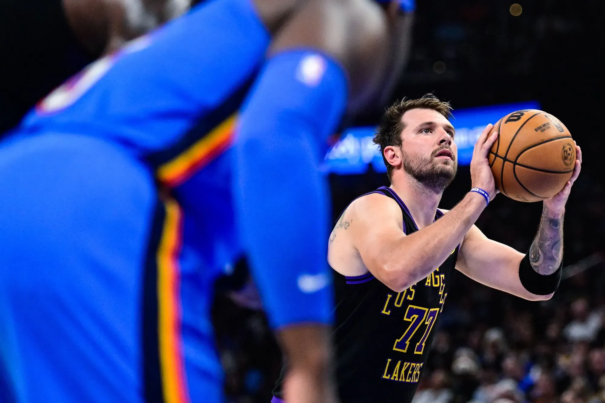 لوكا دونسيتش لاعب لوس أنجلوس ليكرز يستعد لتسديد كرة سلة، بينما يظهر أحد المدافعين في الخلفية، وسط أجواء مباراة حماسية.