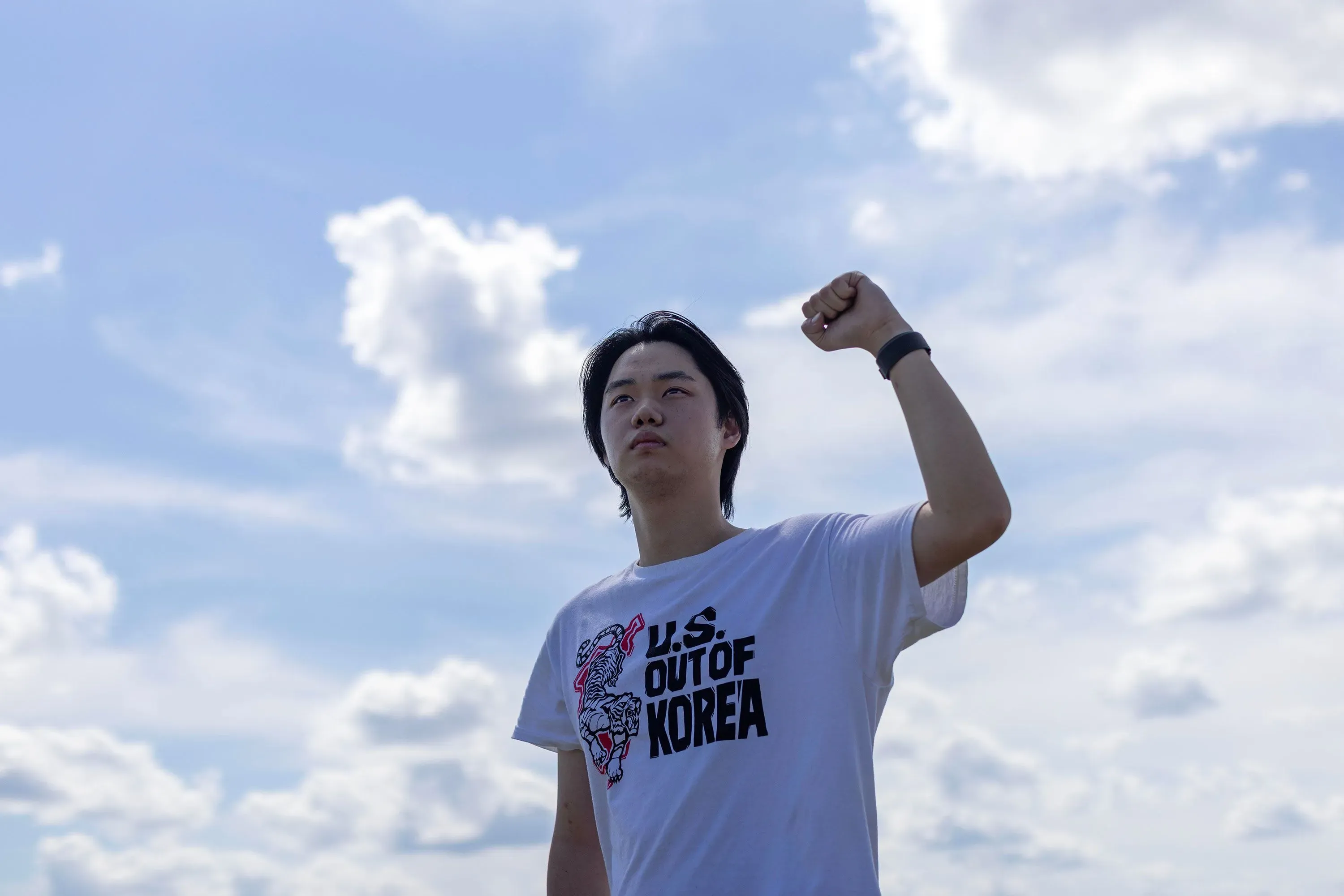 شاب يرفع قبضته في الهواء، مرتديًا قميصًا مكتوبًا عليه "U.S. OUT OF KOREA"، مع سماء زرقاء ملبدة بالغيوم في الخلفية.