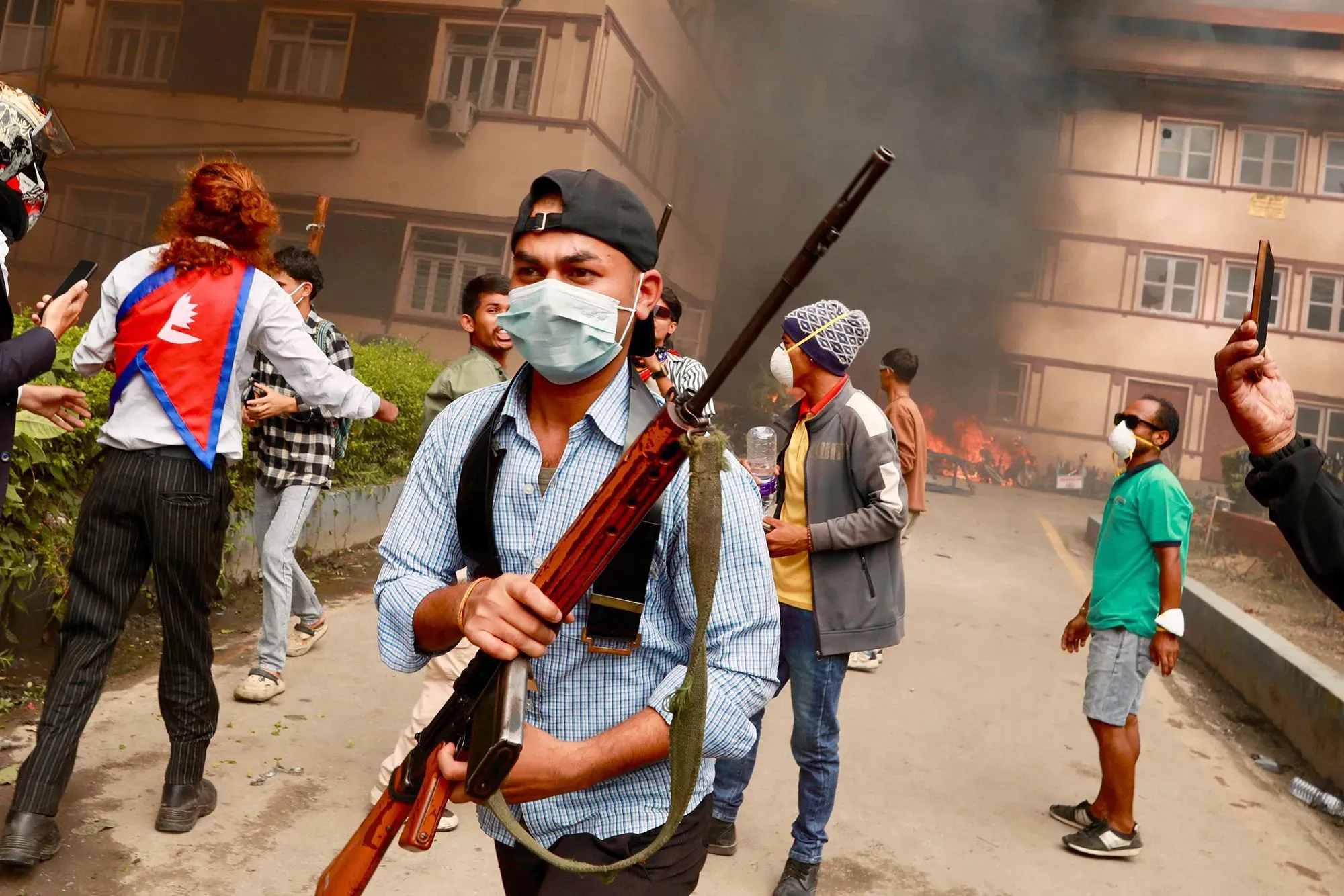 محتجون في نيبال يحملون أسلحة ويتجمعون في موقع احتجاجي، مع تصاعد الدخان من حريق خلفهم، في سياق الاضطرابات السياسية.