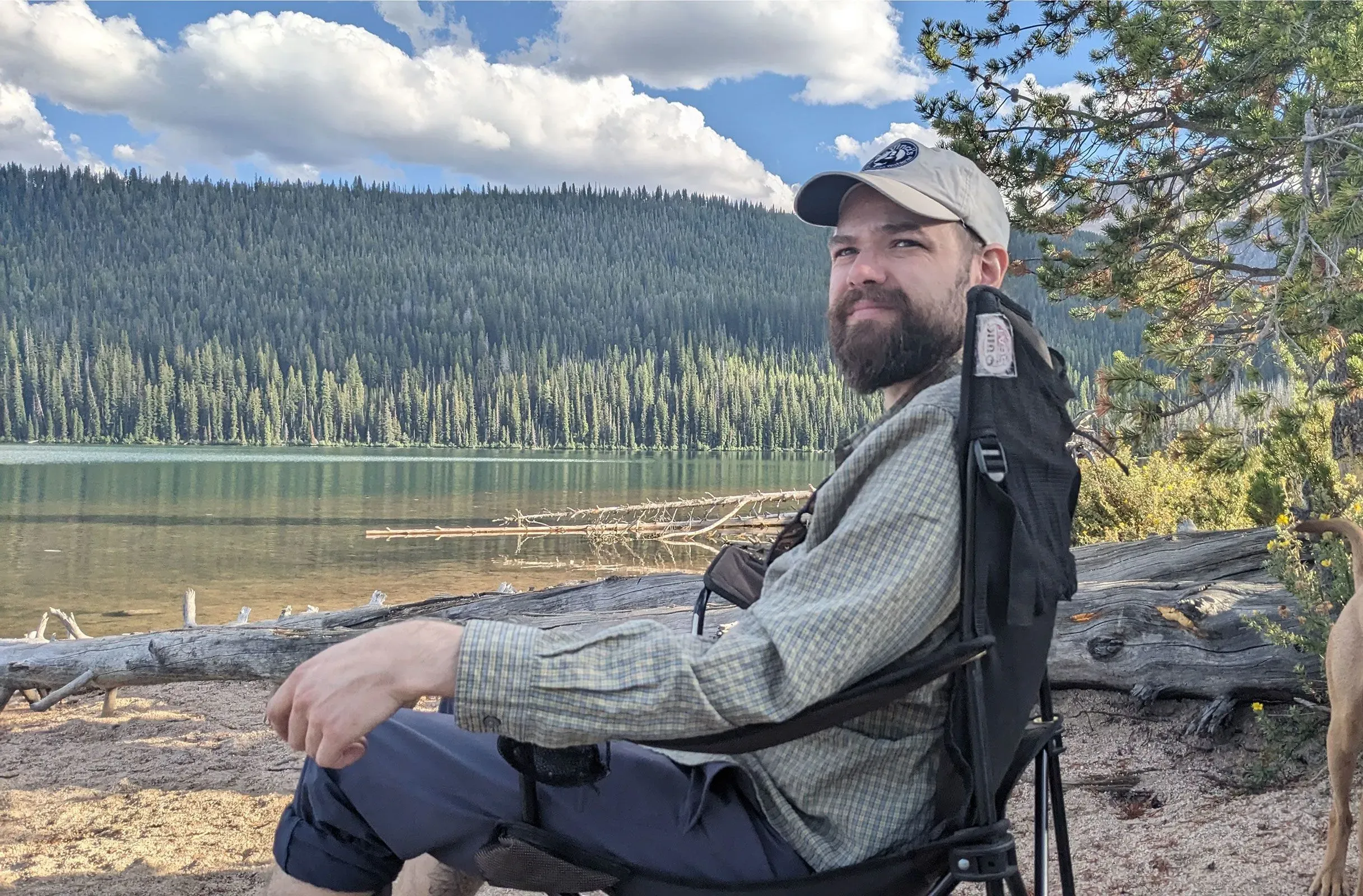 رجل يجلس على كرسي في منطقة طبيعية بجانب بحيرة، محاط بأشجار كثيفة، يعكس شعور الاسترخاء في بيئة هادئة.