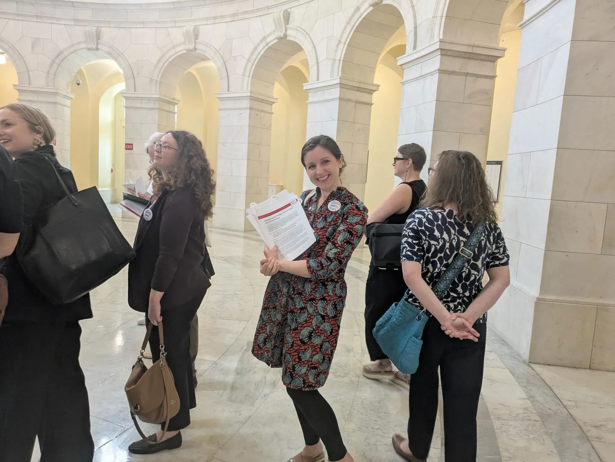 امرأة تحمل أوراقًا في قاعة مبنى حكومي، محاطة بمجموعة من الأشخاص، تعكس تأثير تقليص القوى العاملة الفيدرالية.