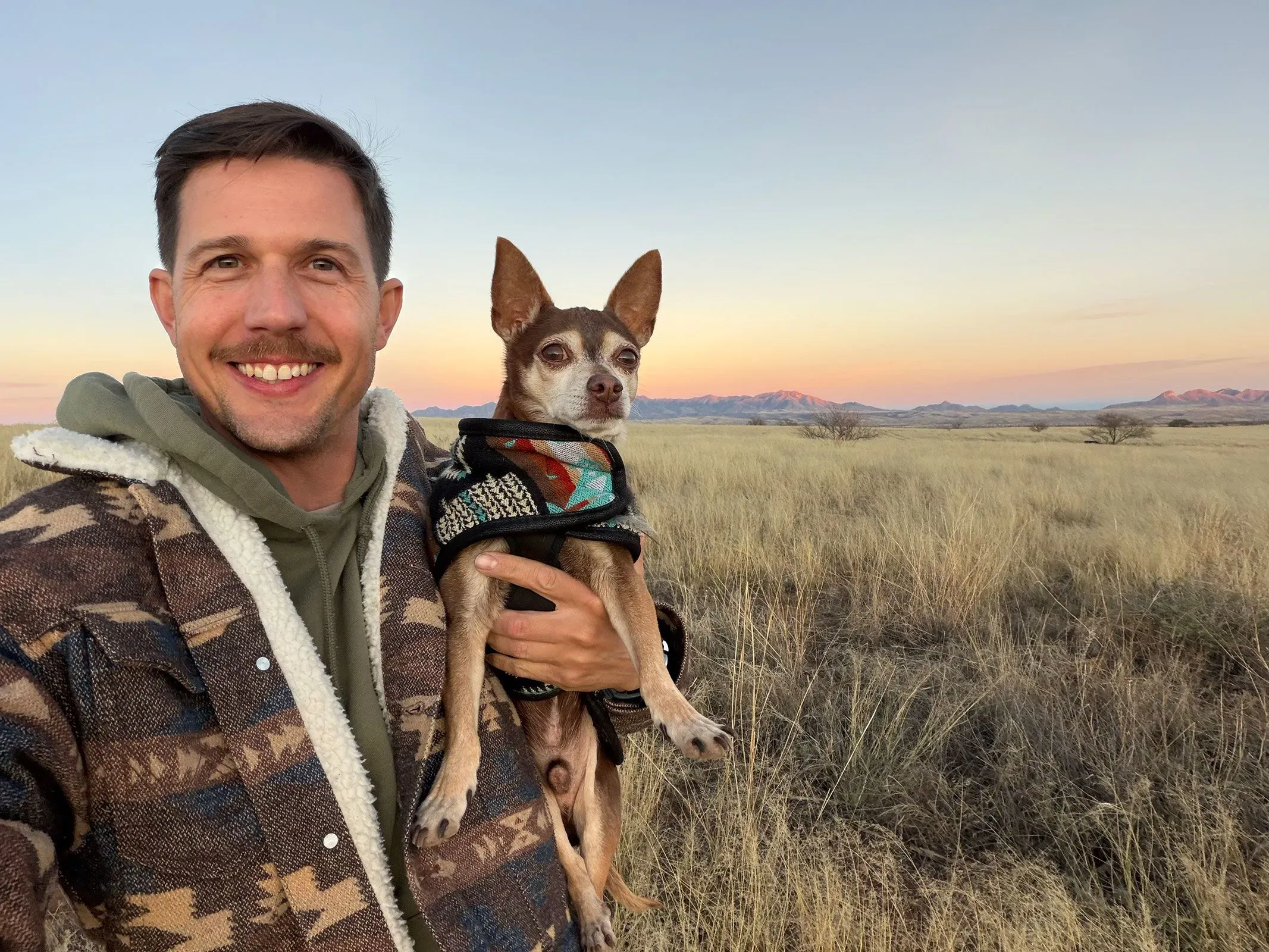 رجل مبتسم يحمل كلبه الصغير في منطقة مفتوحة مع منظر طبيعي خلفه، يعكس شعورًا بالراحة والسكينة.