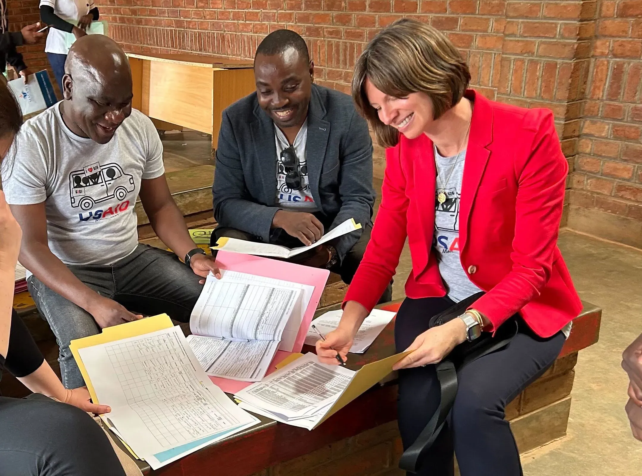 اجتمع مجموعة من الأفراد، بينهم امرأة ترتدي سترة حمراء، حول طاولة، يتبادلون الوثائق والملاحظات المتعلقة بمشاريع الوكالة الأمريكية للتنمية الدولية.