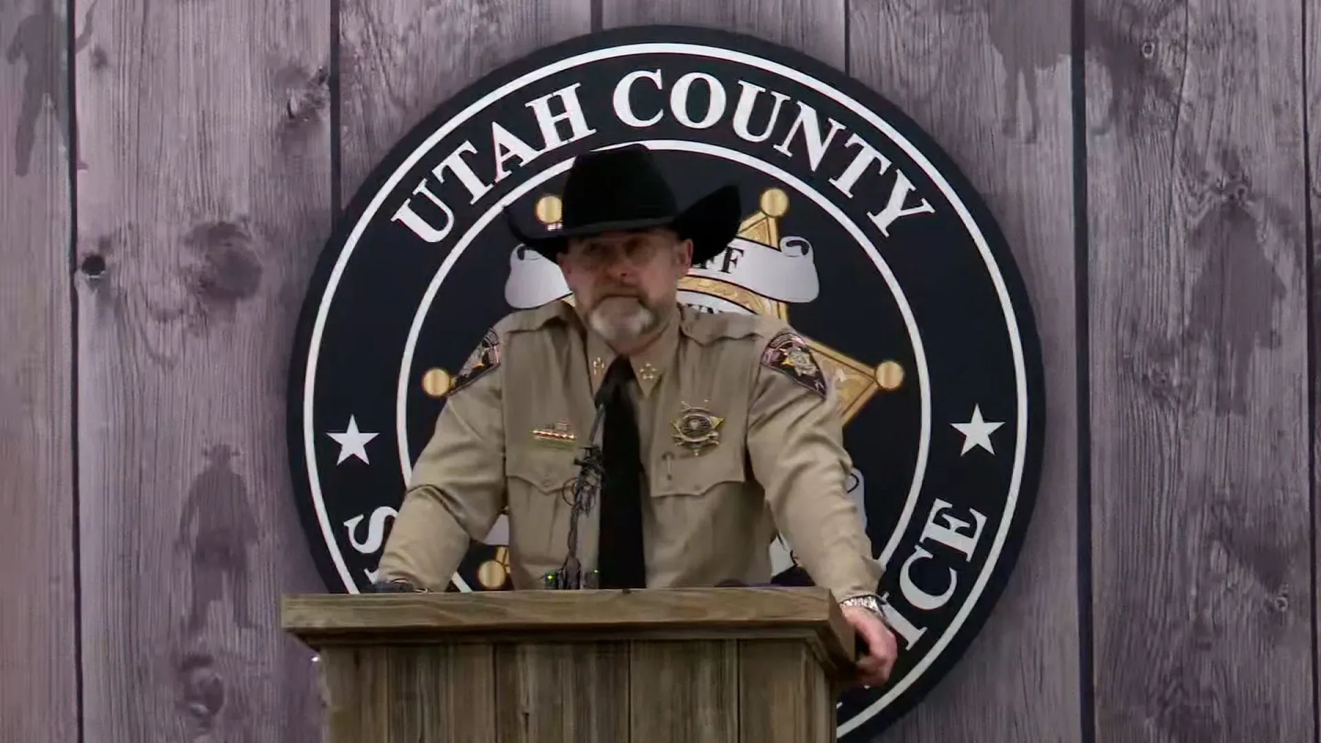 رجل يرتدي زي شرطة يتحدث أمام منصة، مع شعار مكتب شريف مقاطعة يوتا خلفه، خلال مؤتمر صحفي حول قضية لورا آن إيمي.