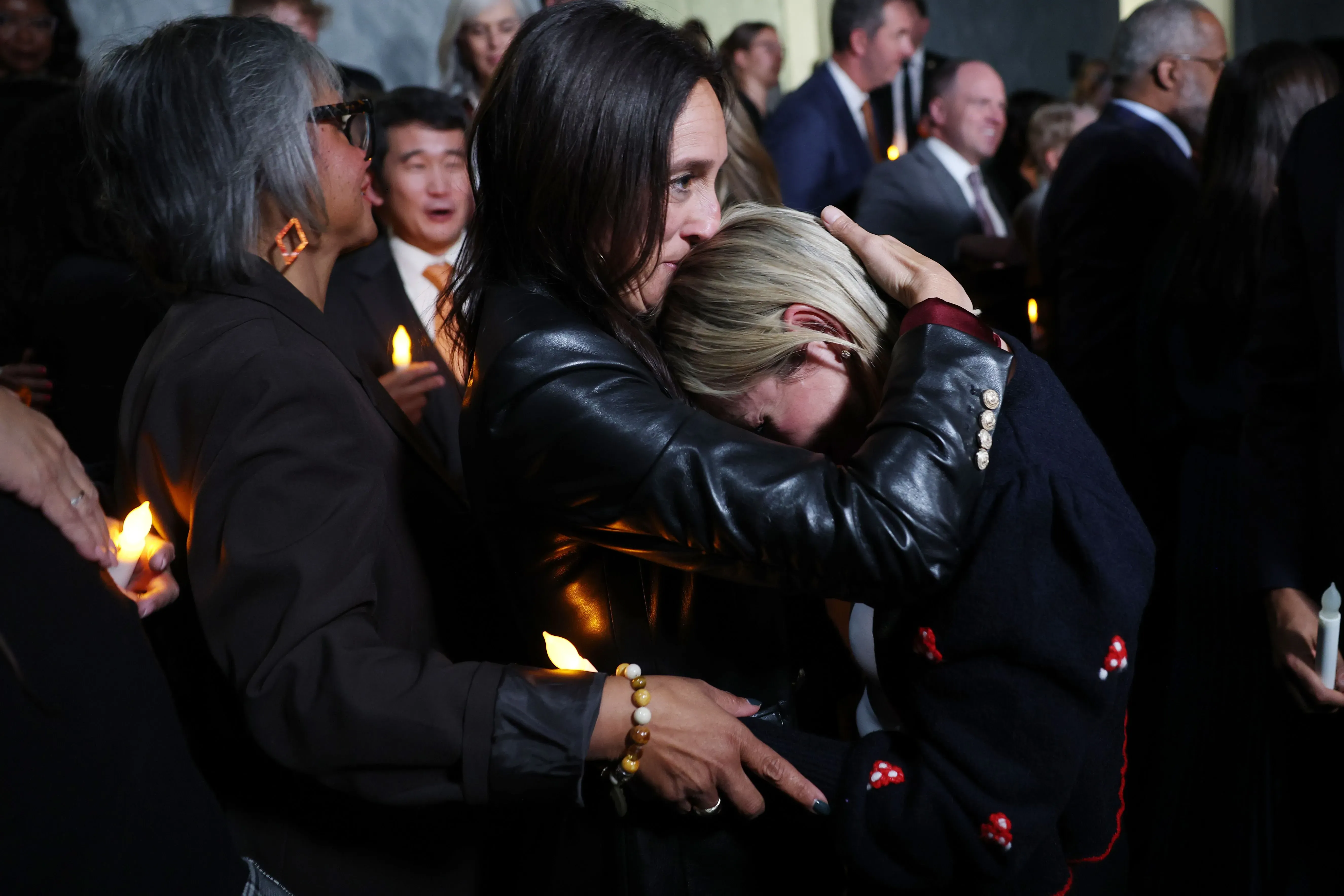 امرأة تعانق أخرى بحزن خلال حدث تأبيني، مع وجود شموع مضاءة في أيدٍ أخرى، تعكس مشاعر الألم والقلق بشأن ضحايا جيفري إبشتاين.