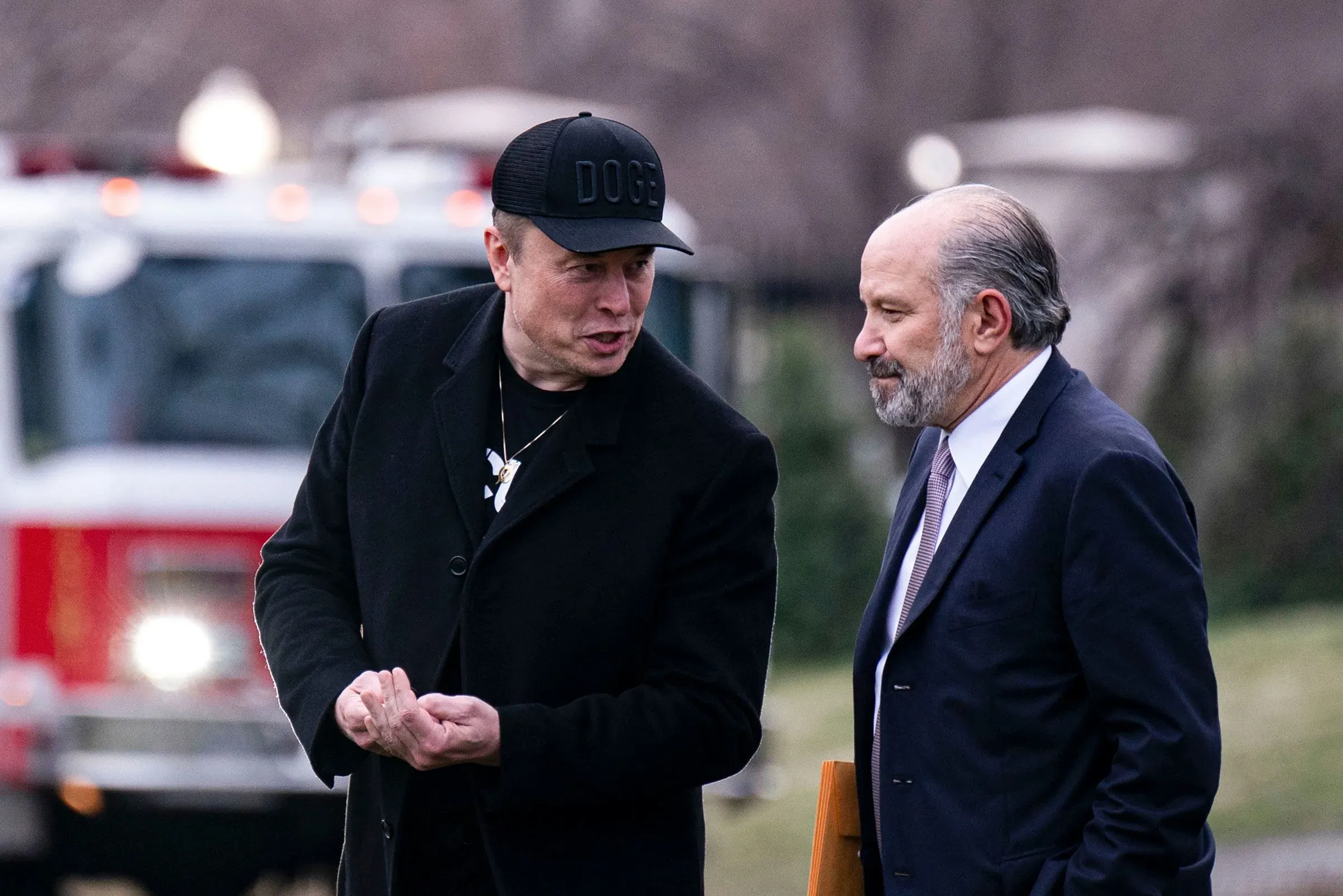 إيلون ماسك يتحدث مع مسؤول حكومي، بينما تظهر سيارة إطفاء في الخلفية، في سياق مناقشات حول قضايا قانونية معقدة.