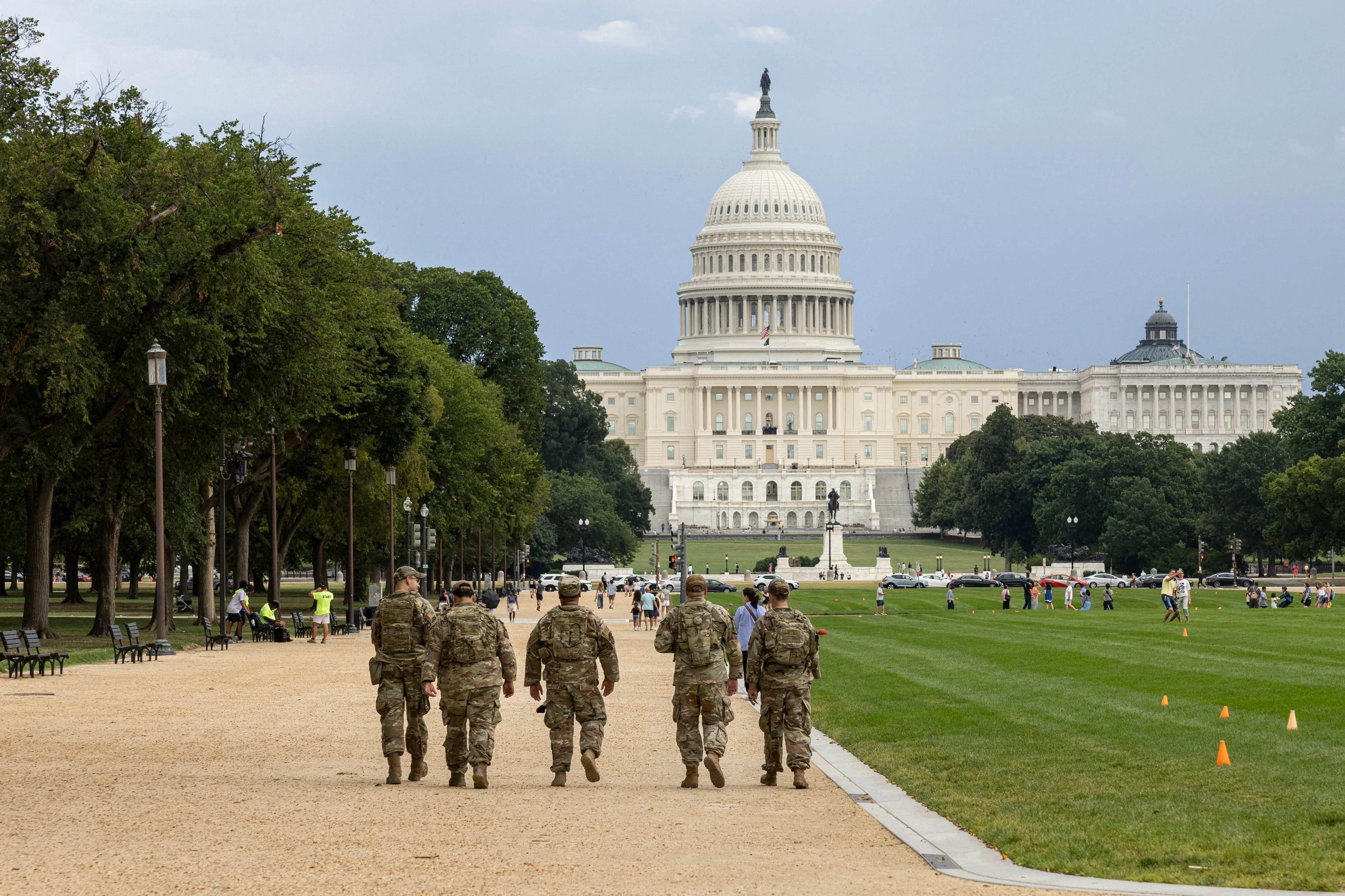 جنود من الحرس الوطني يسيرون في منطقة مفتوحة بالقرب من مبنى الكابيتول، وسط أشخاص ومرافق خضراء، في إطار جهود الأمن العام.