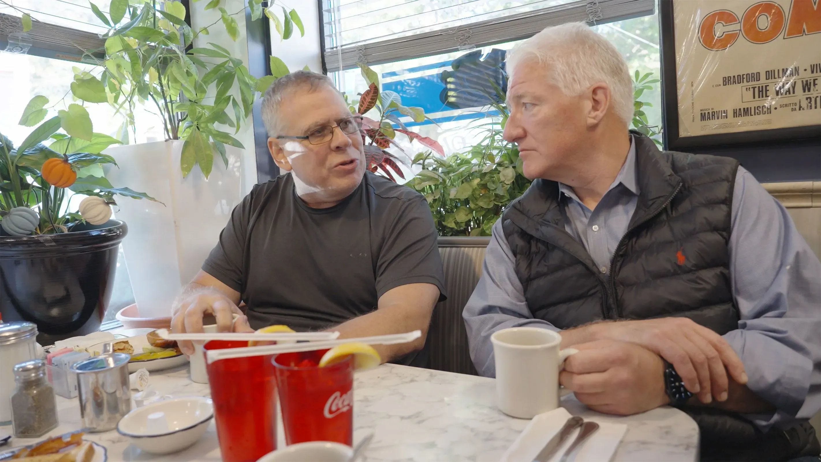 محادثة بين رجلين في مطعم في بروكلين، مع نباتات في الخلفية، حيث يناقشان قضايا الانتخابات المحلية وتأثيرها.