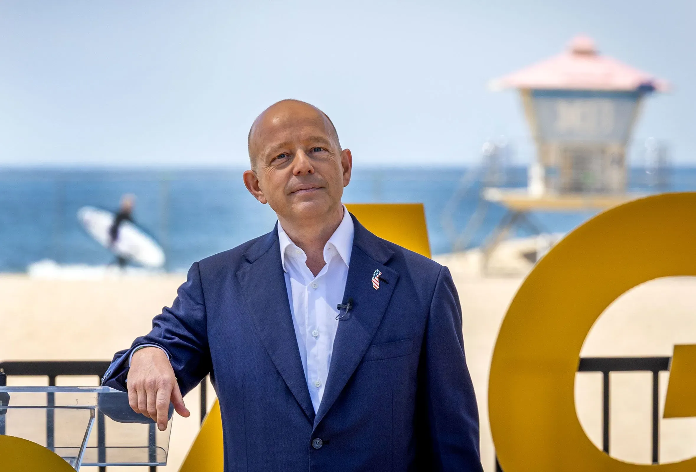 حاكم ولاية كاليفورنيا جافين نيوسوم يقف أمام شاطئ مع لافتة كبيرة، حيث يتحدث عن خطته لإعادة رسم خرائط الكونغرس وسط معارضة.