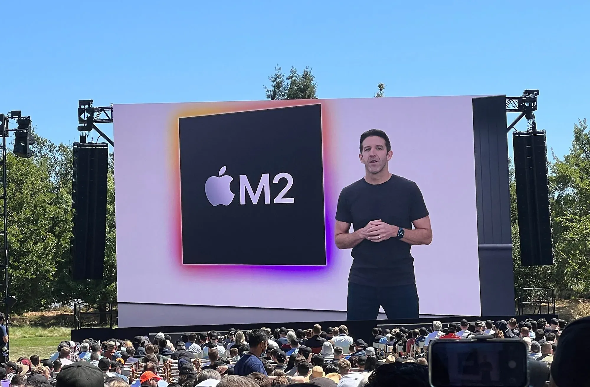 جون تيرنوس يتحدث في مؤتمر عن شريحة Apple M2، مع جمهور كبير في الخلفية، مما يعكس أهمية الابتكار في عهد القيادة الجديد.