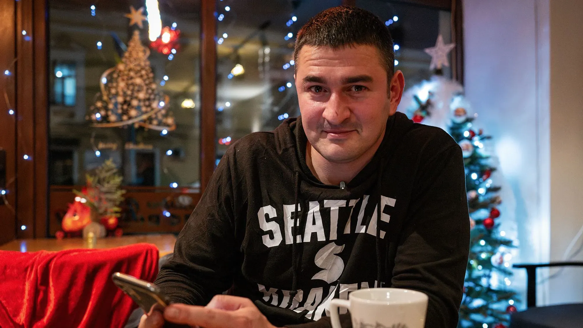 فيكتور بينخاسوف، شاب أوكراني، يجلس مبتسمًا في مقهى برومانية، محاطًا بزينة عيد الميلاد، بعد فراره من أوكرانيا.