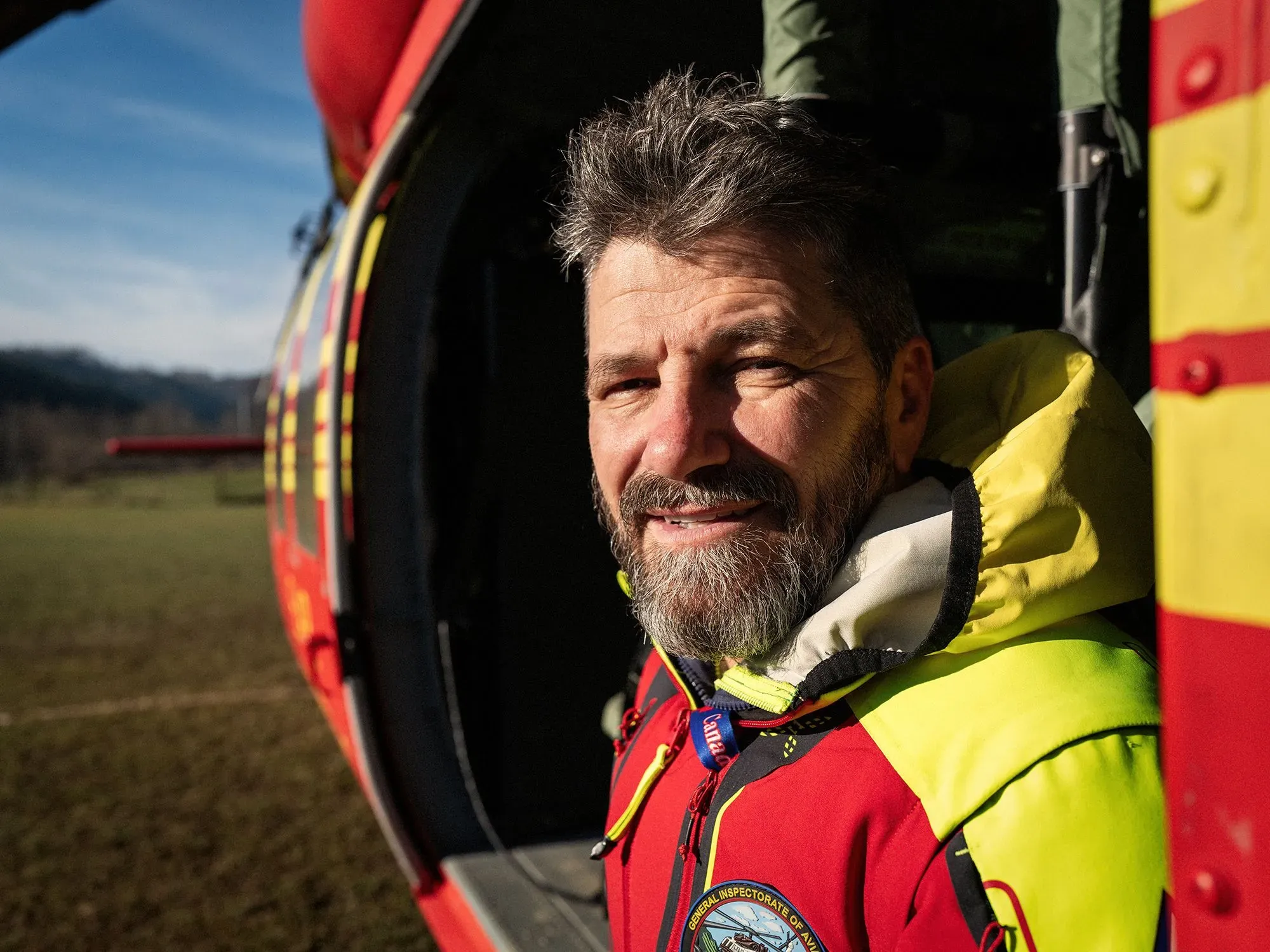 رجل يرتدي زي الإنقاذ الأحمر يقف بجوار مروحية، مبتسمًا، في منطقة جبلية، مع خلفية طبيعية واضحة، يعكس جهود الإنقاذ للأوكرانيين الفارين.