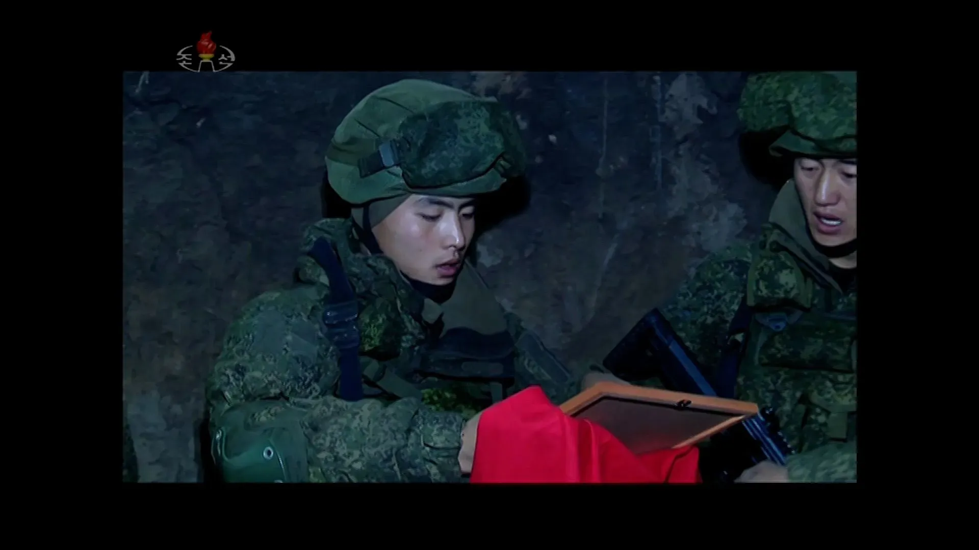 جنود يرتدون زيًا عسكريًا في موقع مظلم، يتبادلون الحديث حول وثيقة حمراء، مما يعكس أجواء الحرب والجهود العسكرية لكوريا الشمالية.