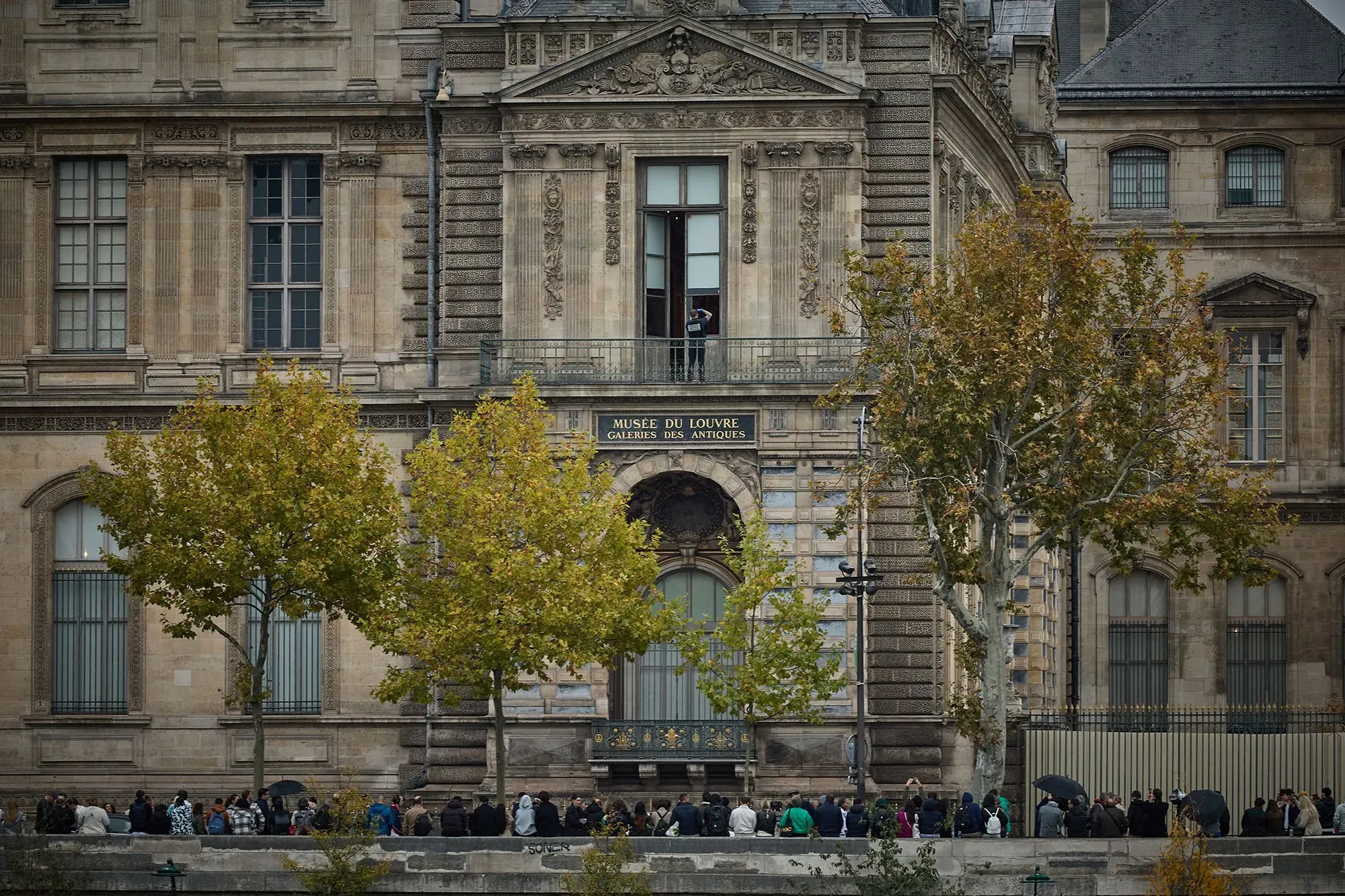 طابور طويل من الزوار أمام متحف اللوفر في باريس، مع تفاصيل معمارية مميزة للمتحف وأشجار خريفية في المقدمة.
