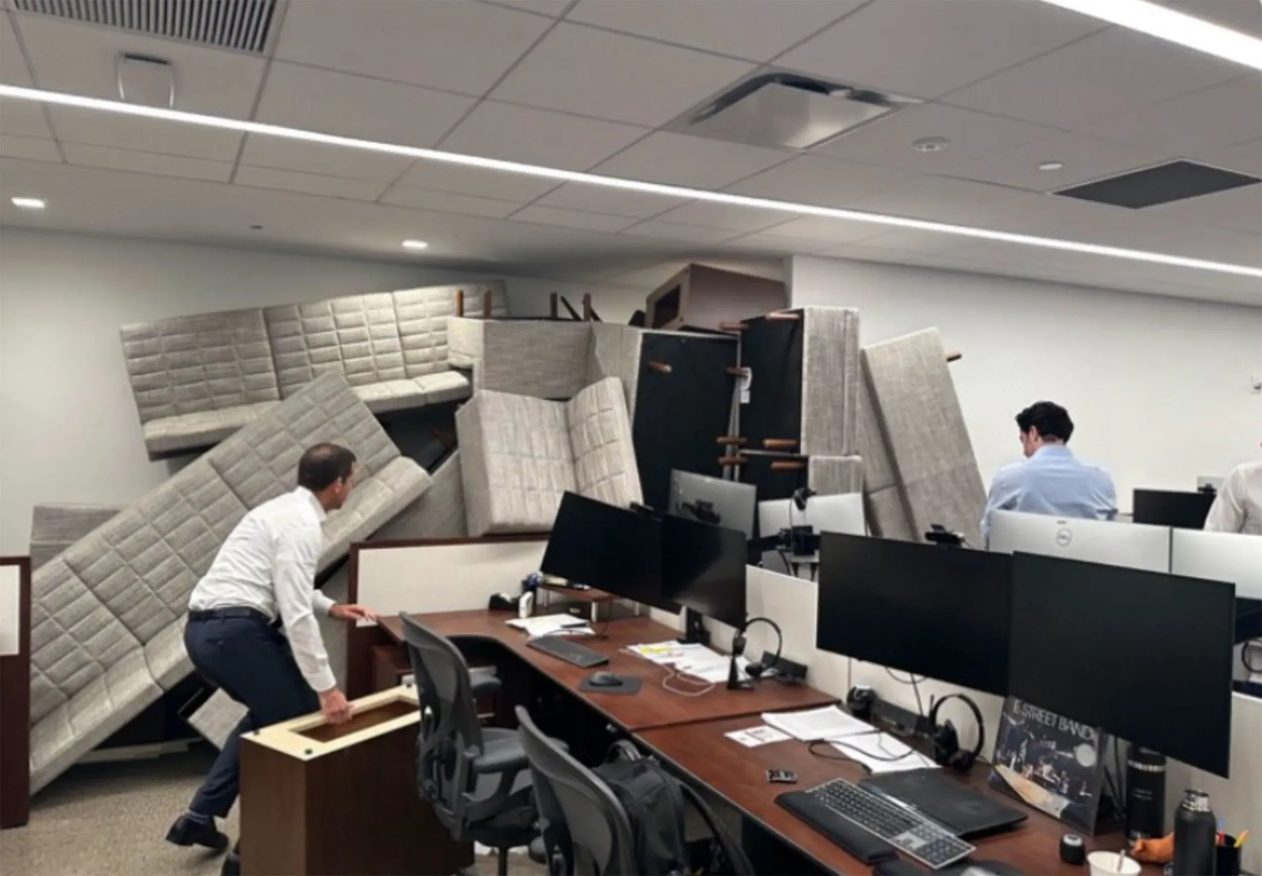 موظفان يحاولان إبعاد الأثاث المتراكم في مكتب بعد حادث إطلاق نار، مع وجود شاشات كمبيوتر وأدوات مكتبية في الخلفية.