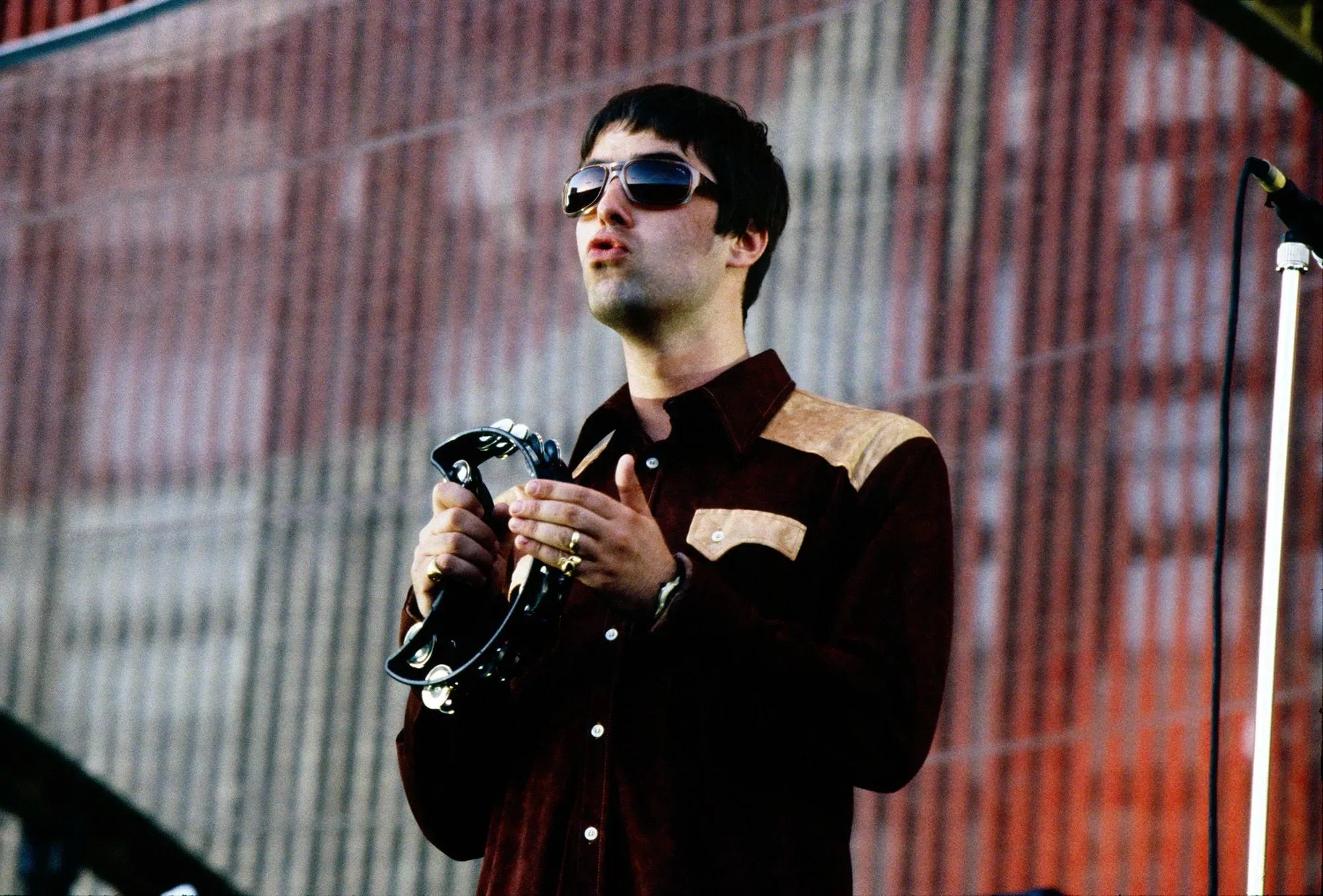 ليام غالاغر يؤدي في عرض حي، ممسكًا بآلة التامبورين، يعبر عن طاقته وشغفه بالموسيقى، في لحظة من تاريخ فرقة أويسس.