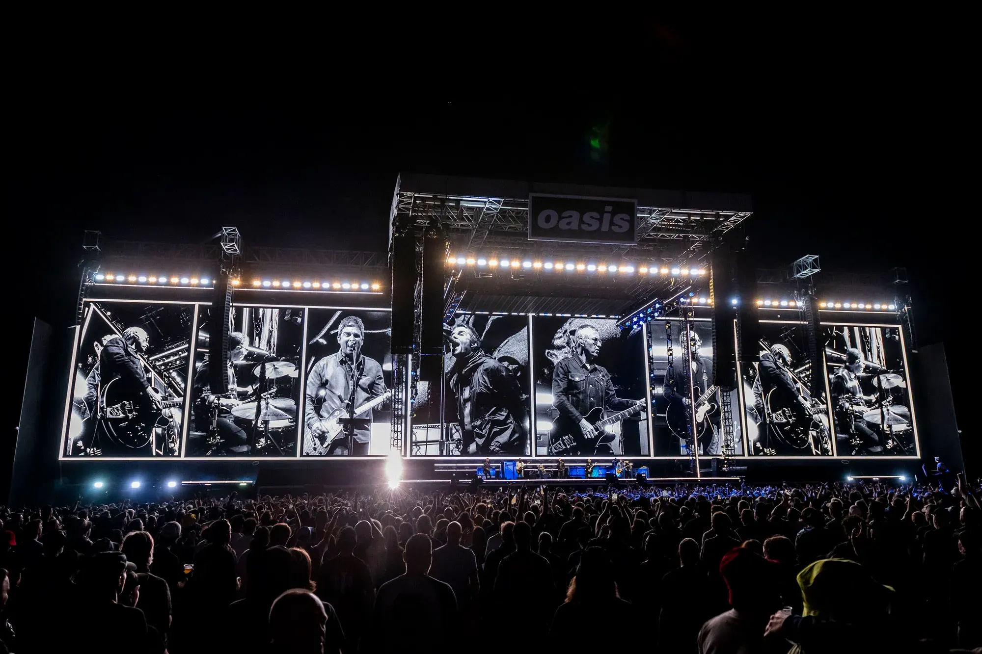 عرض فرقة أويسس في ملعب كبير، مع أضواء ساطعة وجمهور ضخم، يعكس عودتهم المثيرة إلى الساحة الموسيقية بعد سنوات من الانقسام.