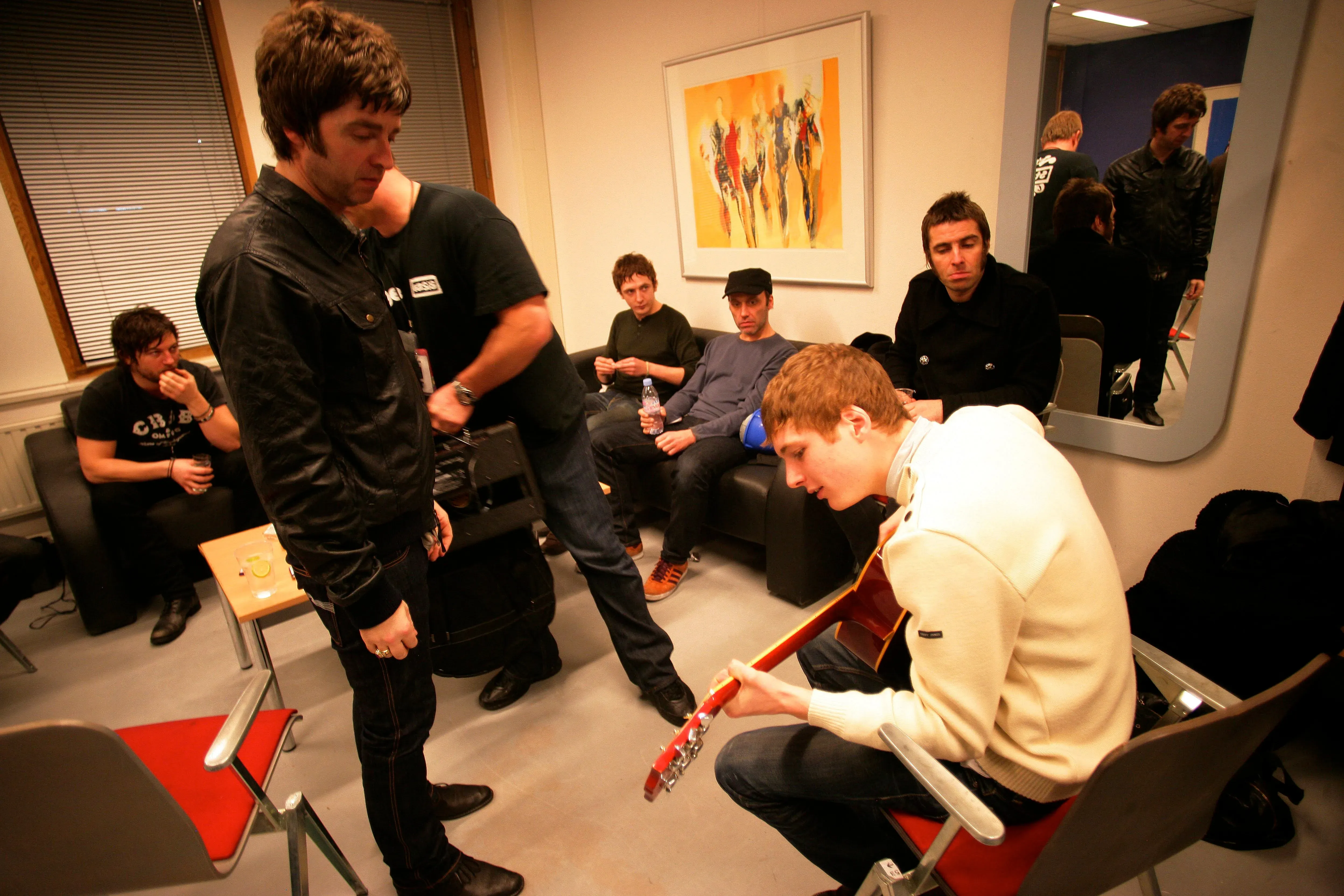 مجموعة من الأشخاص في غرفة خلف الكواليس، حيث يعزف أحدهم على الجيتار بينما يتحدث آخرون، مما يعكس أجواء فرقة أويسس قبل عرضهم.