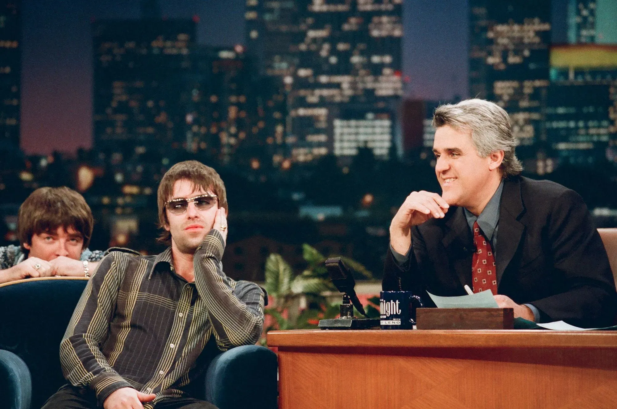 ليام غالاغر يجلس في برنامج حواري، مرتديًا نظارات شمسية، مع خلفية لمدينة لوس أنجلوس، بينما يظهر شقيقه نويل خلفه.