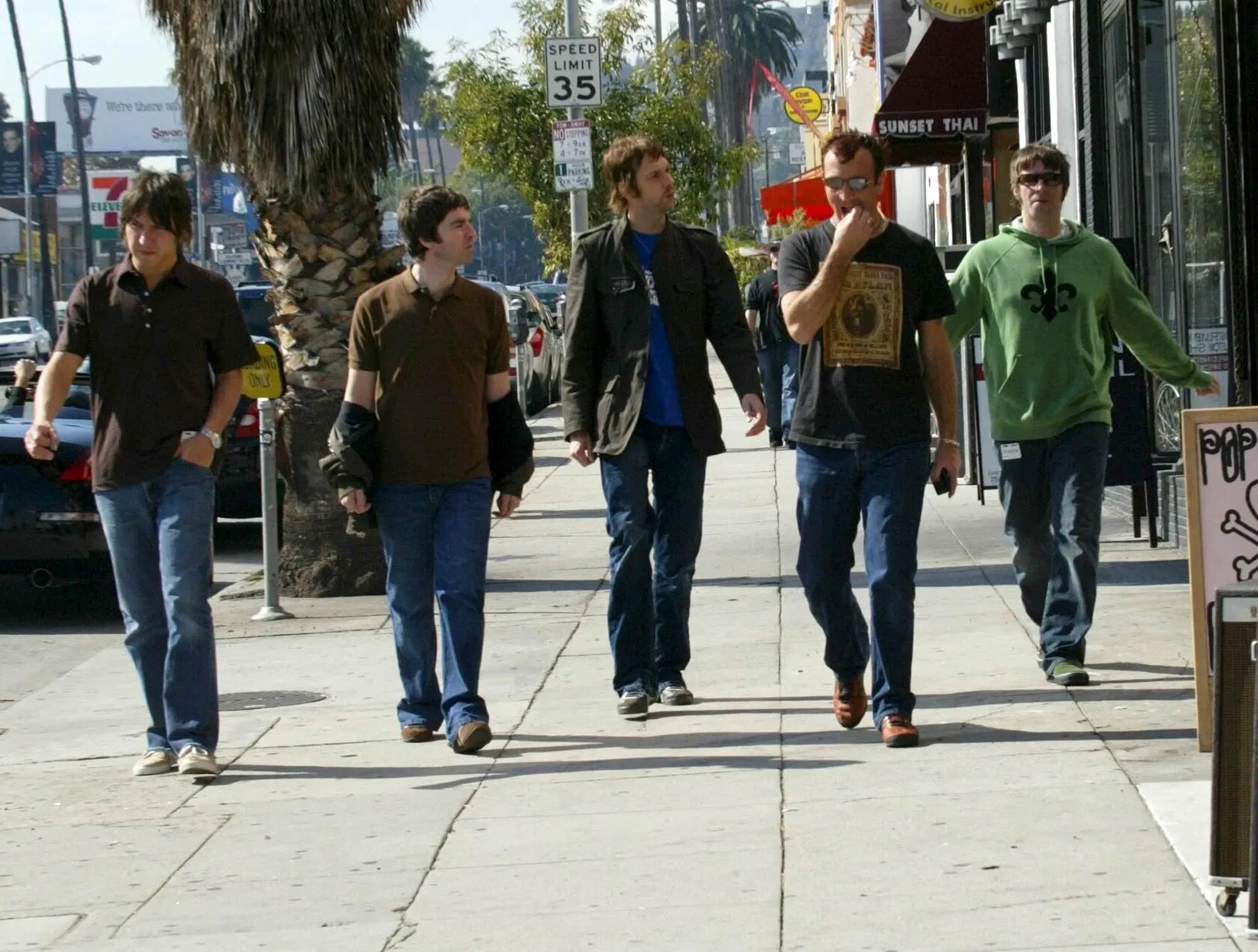 مجموعة من الرجال يسيرون على رصيف في حي حضري، مع أشجار النخيل والمحال التجارية في الخلفية، يعكسون أجواء ثقافة الشارع.