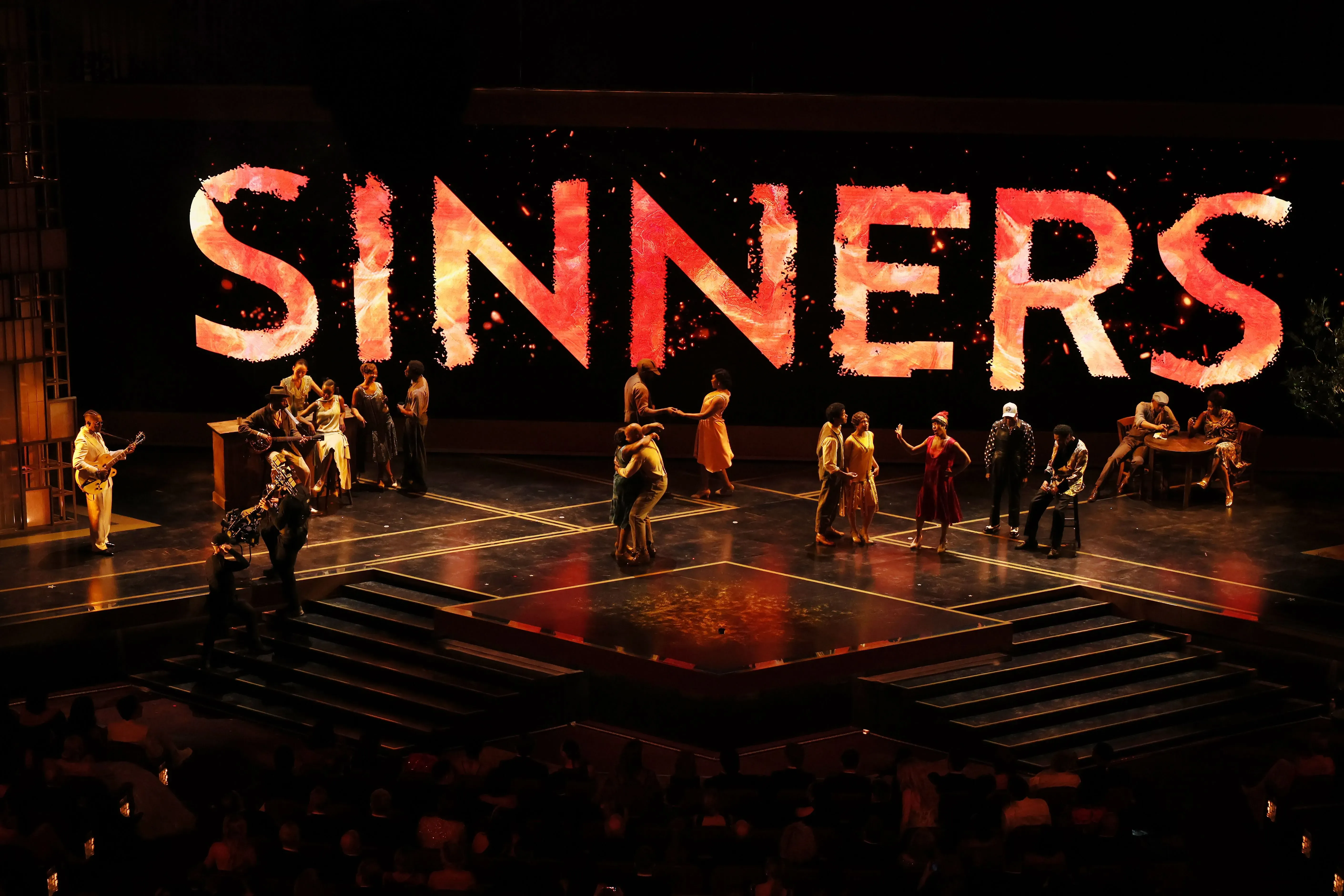 عرض موسيقي حي من فيلم "المذنبون"، مع خلفية تحمل كلمة "SINNERS" مضاءة، وراقصون يؤدون على المسرح.