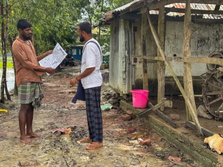 حسن بارفيز يناقش محتوى صحيفة "أندهارمانيك" مع رجل آخر في قرية غرب سوناتالا، بجوار منزل بسيط ومحيط طبيعي.