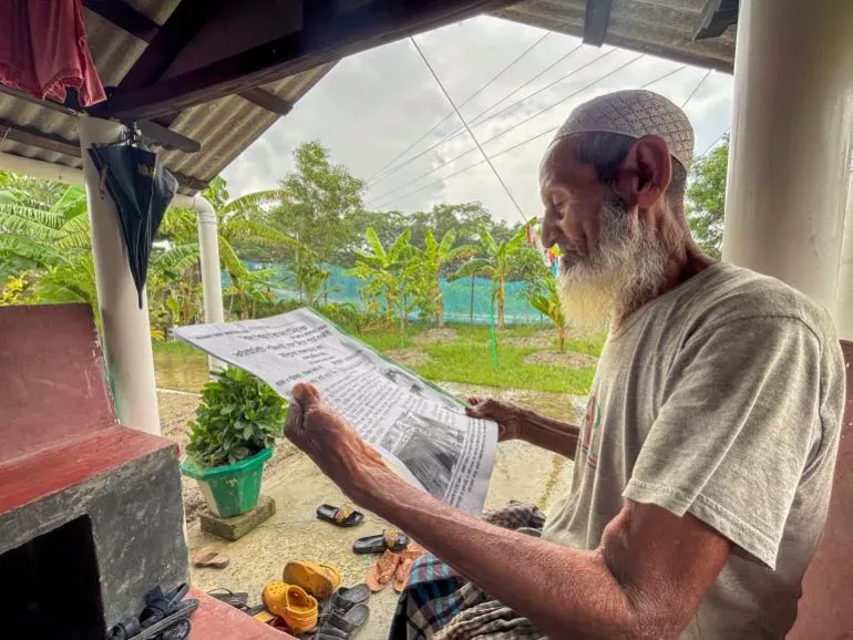 رجل مسن يجلس في فناء منزله، يقرأ صحيفة مكتوبة بخط اليد، محاطًا بأشجار ومزارع. تعكس الصورة أهمية الصحافة المجتمعية في قريته.