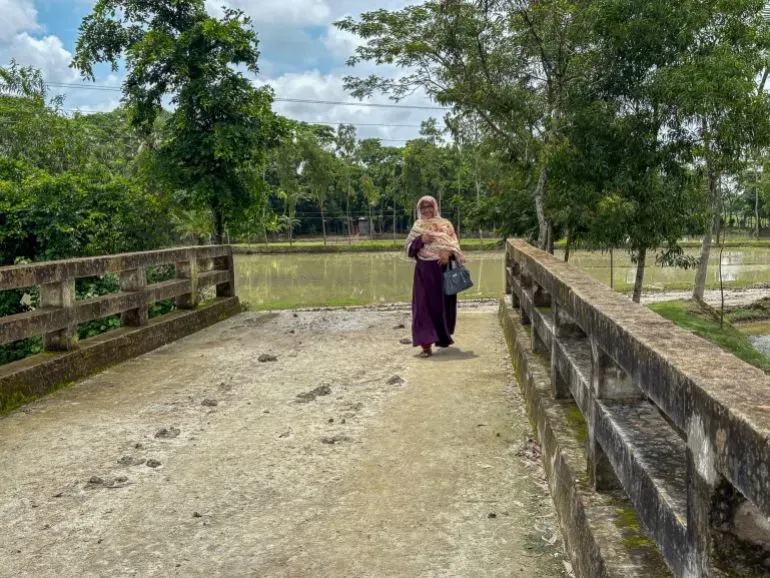 امرأة ترتدي عباءة وتحتضن طفلًا، تسير على جسر ضيق وسط الطبيعة الخضراء في غرب سوناتالا، بنغلاديش.