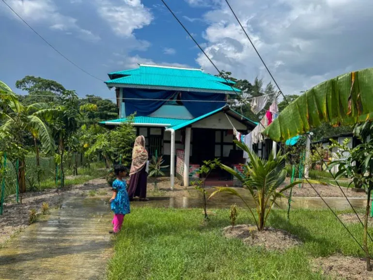 منزل حسن بارفيز ذو السقف الأزرق المحاط بالخضرة والأشجار، مع امرأة ترتدي ملابس تقليدية وطفلة تلعب في الحديقة.