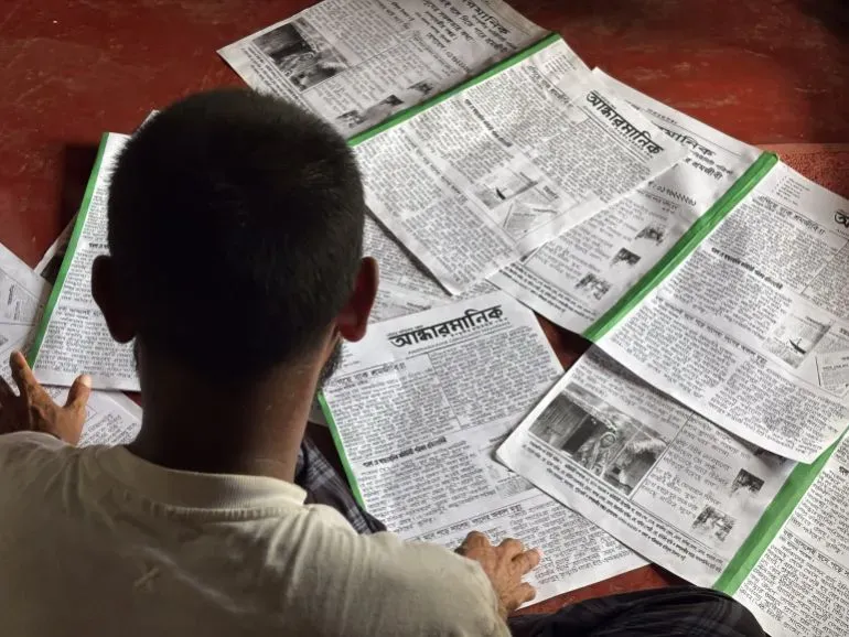 شخص يجلس على الأرض ويقرأ صحيفة "أندهارمانيك" المجتمعية المكتوبة بخط اليد، محاطًا بعدد من النسخ المفتوحة.