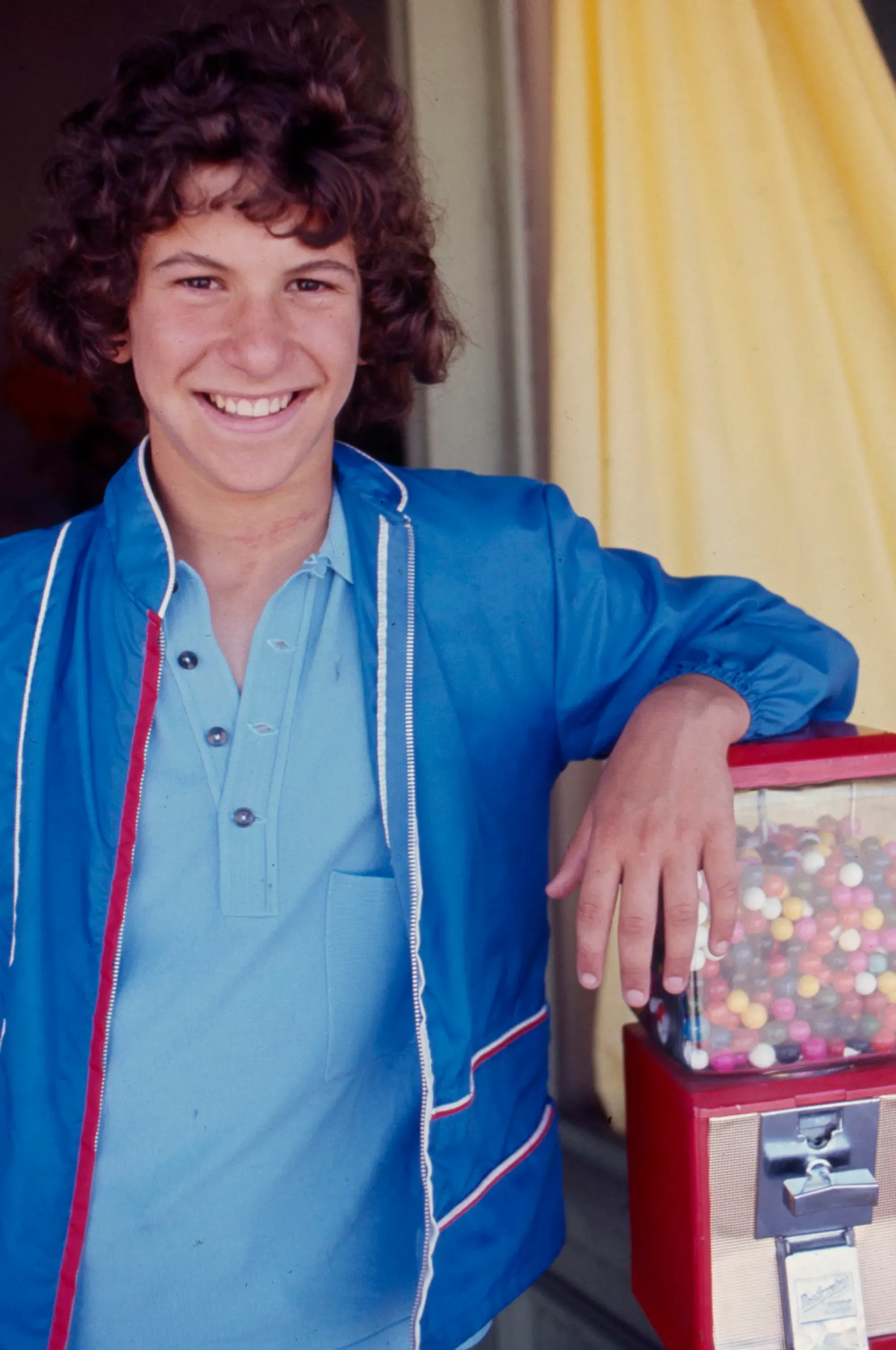 شاب مبتسم يرتدي سترة زرقاء، يقف بجانب آلة لبيع الحلوى، تعكس لحظة من الطفولة السعيدة.