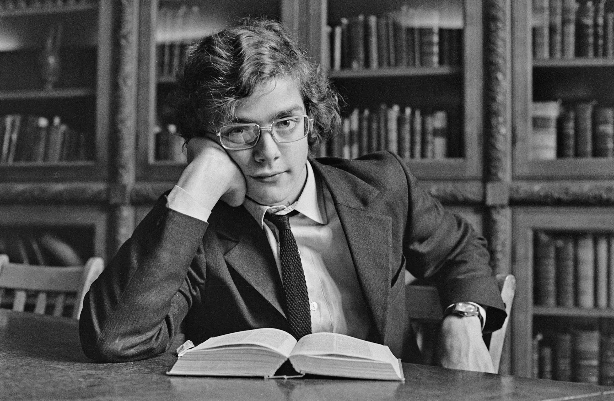 شاب يرتدي بدلة ويضع يده على خده، يجلس أمام كتاب مفتوح في مكتبة، تعكس الصورة أجواء الدراسة والتفكير.