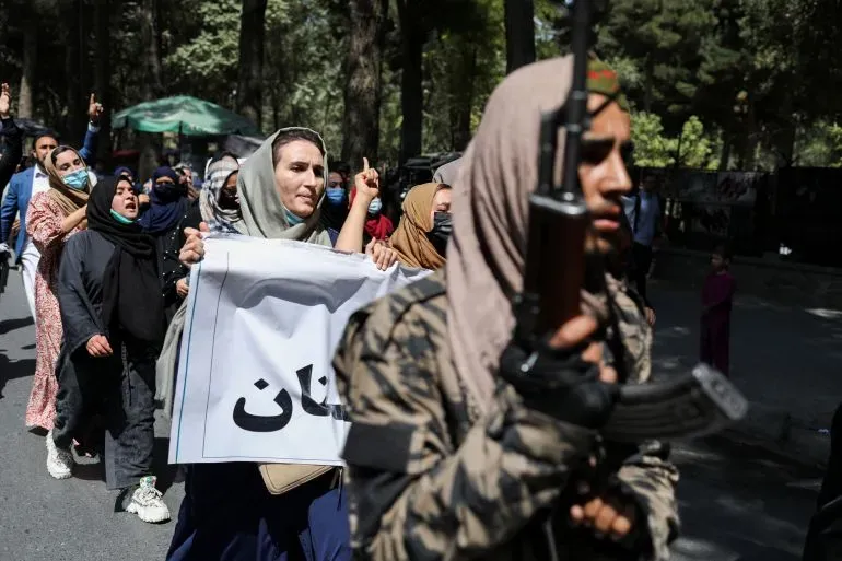 نساء يحملن لافتة خلال احتجاج في كابول، بينما يراقب مسلح من طالبان الوضع، مما يعكس التوترات السياسية في أفغانستان.