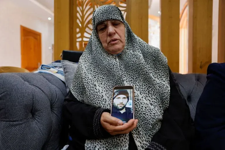 امرأة مسنّة ترتدي حجابًا، تجلس على أريكة وتظهر صورة لابنها الراحل، تعبيرها يعكس الحزن بعد مقتل الفلسطيني خميس عياد في هجوم للمستوطنين.